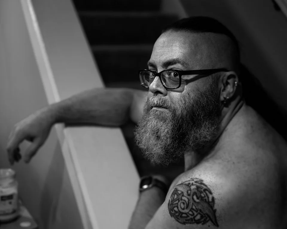 A black and white portrait of a shirtless man with glasses, a beard, and a tattoo on his shoulder, sitting and leaning on a surface, looking to the side.