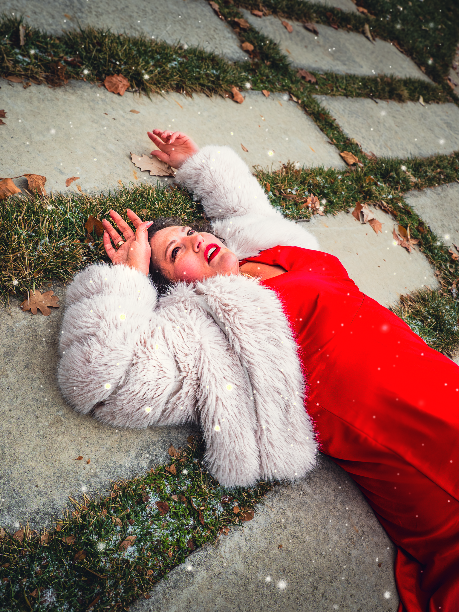 Woman in a red dress and white faux fur coat lying on concrete sidewalk next to grass and fallen leaves, looking upwards.