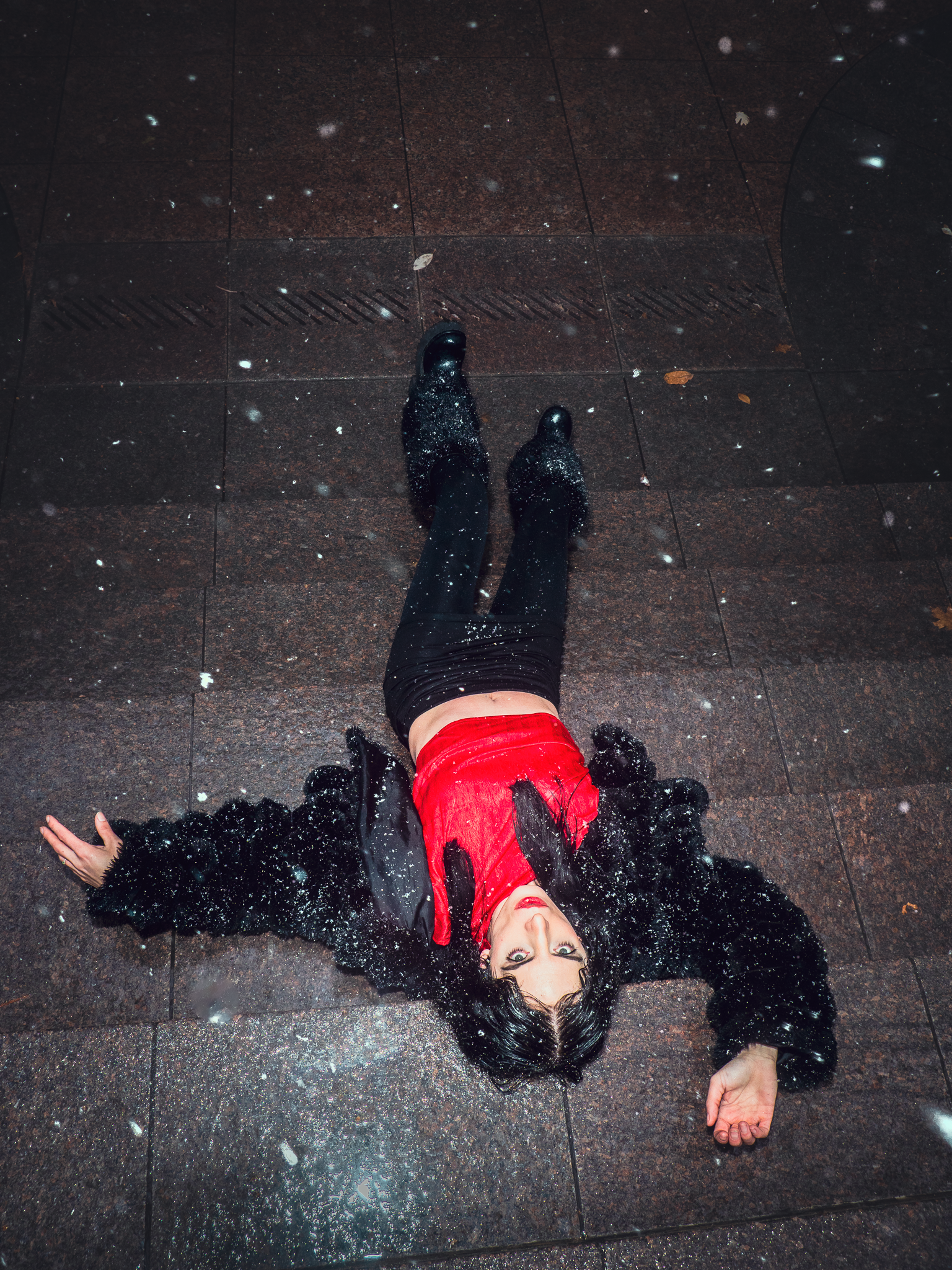 A woman lies on the wet pavement in the rain, wearing a black furry coat, red top, and black pants, with raindrops on her clothes and face, and a neutral expression.