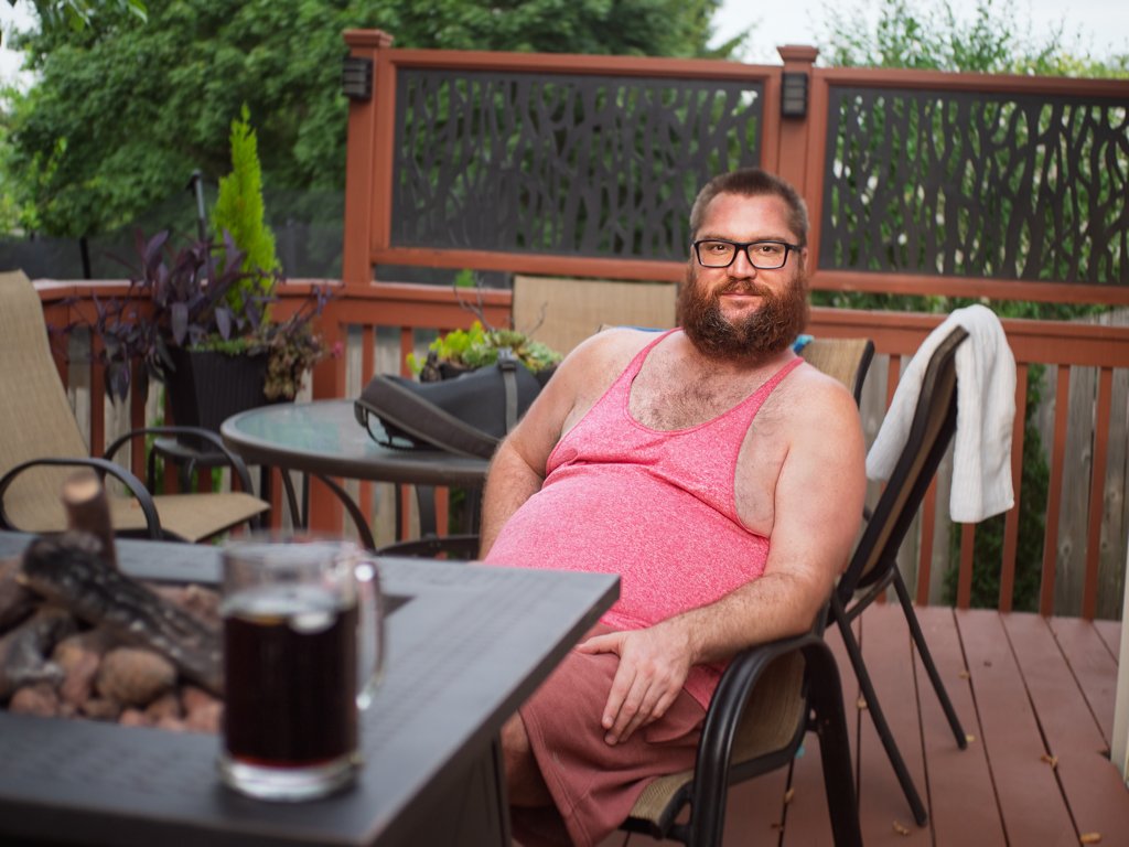 A man with glasses and a beard, wearing a pink tank top and shorts, sitting on a patio chair on an outdoor deck. There is a table with a drink and some rocks in the foreground, and potted plants and a wooden fence in the background.