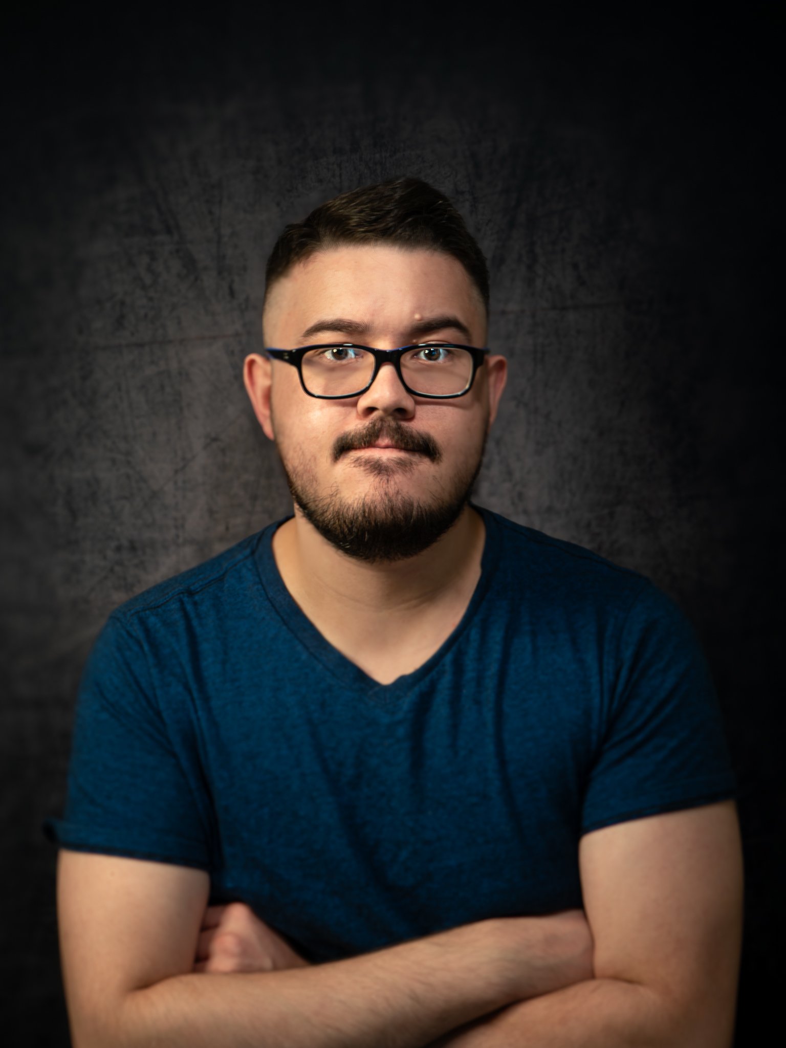 A young man with short dark hair, glasses, and a beard, wearing a blue t-shirt, standing with arms crossed against a dark textured background.