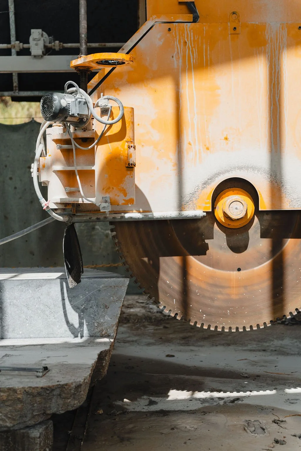 Close-up of a large orange masonry saw with a circular blade cutting concrete, outdoors with sunlight and shadow.