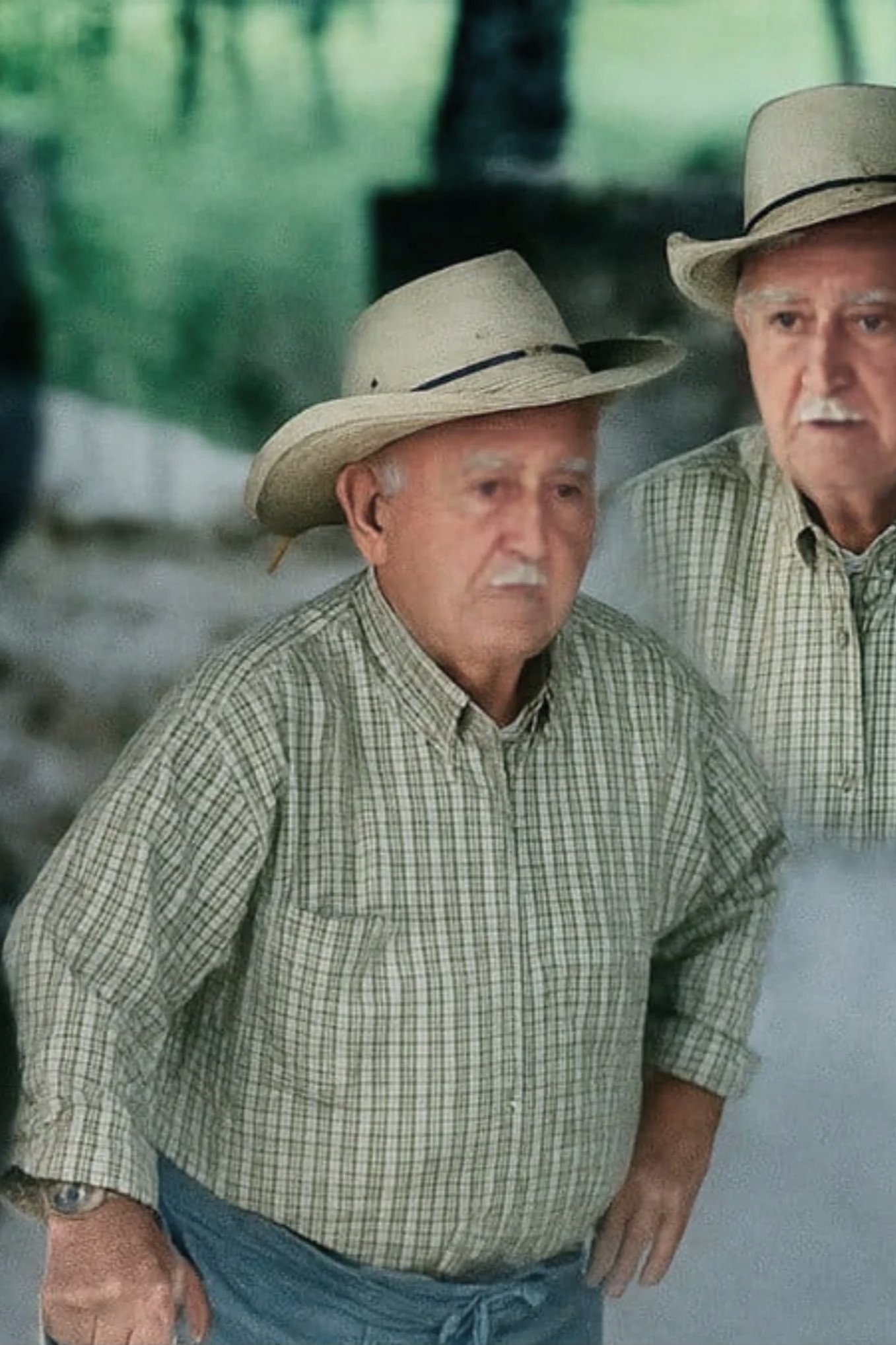 Two elderly men in plaid shirts and straw hats standing outdoors, looking in the same direction. Heritage of limestone knowledge.