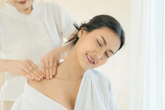 A woman receiving a neck and shoulder massage from a therapist in a bright, clean room.