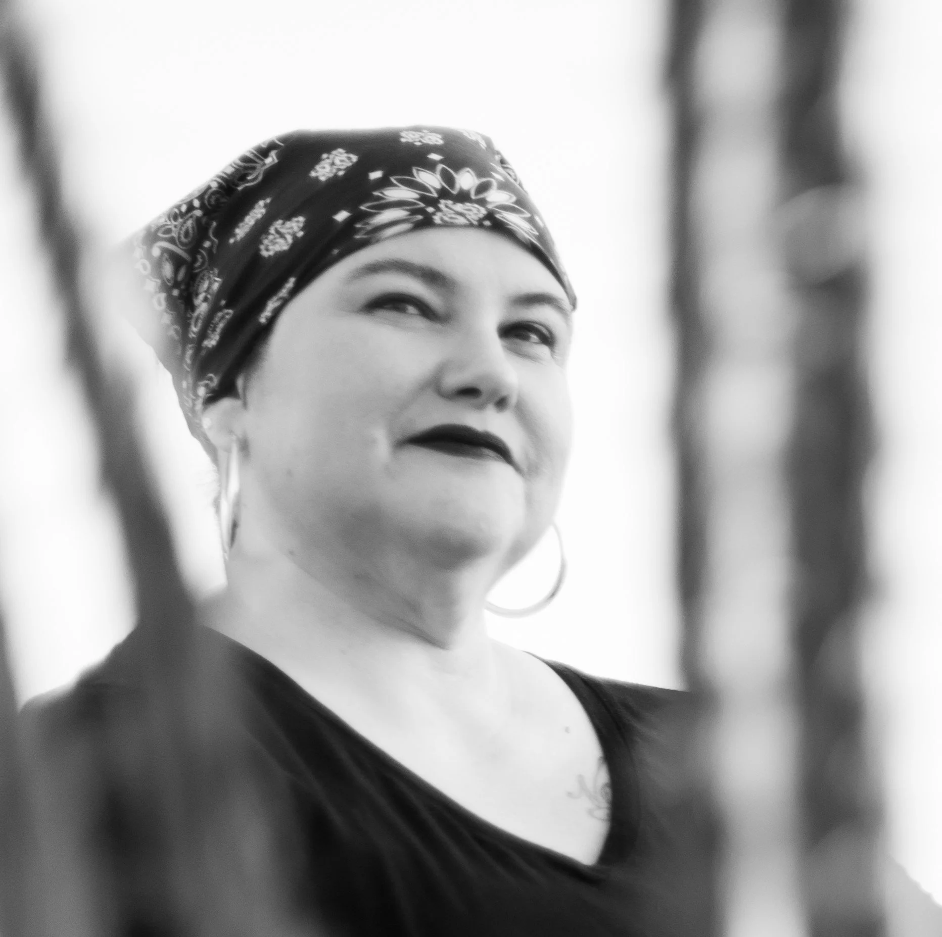 B & W image. Naomi Ortiz, light-skinned Mestize using a mobility scooter, sits behind stalks of an ocotillo, long & narrow limbs with thorns. They wear a bandanna, a V-neck shirt, with silver hoop earrings, & dark lipstick. Photo Credit: Jade Beall.