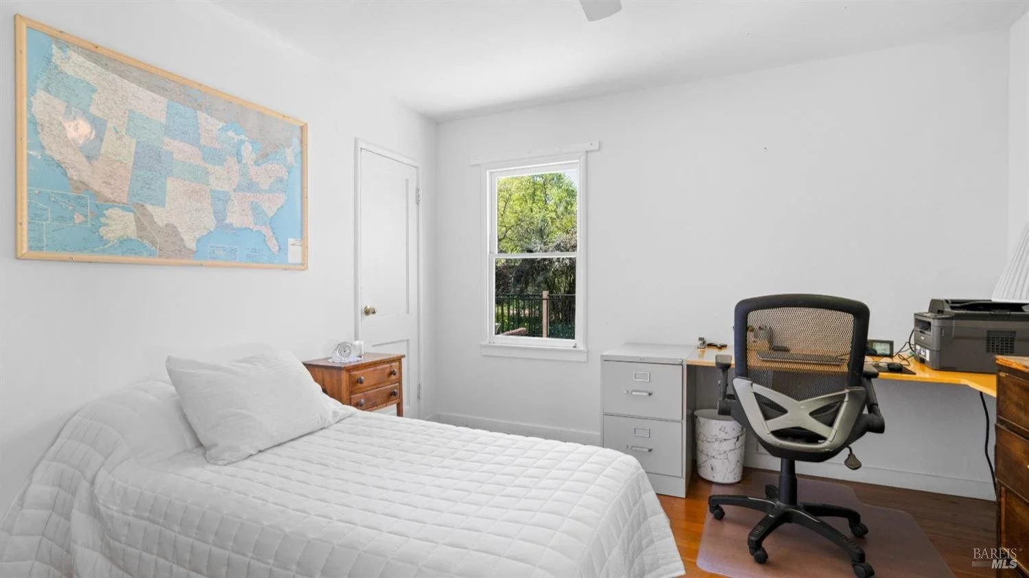 A small, minimalist bedroom with a white bed, wooden dresser, and a desk with an office chair. There is a window with greenery outside and a map of the United States on the wall.