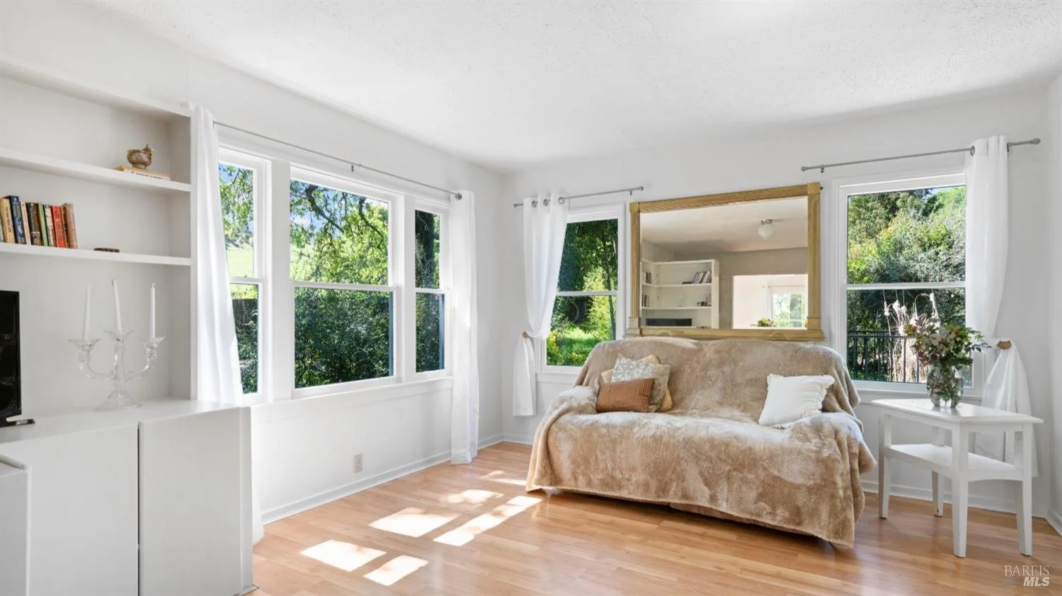 Bright living room with large windows, white walls, wooden floor, beige couch with pillows, white side table with a vase of flowers, and a mirror on the wall.