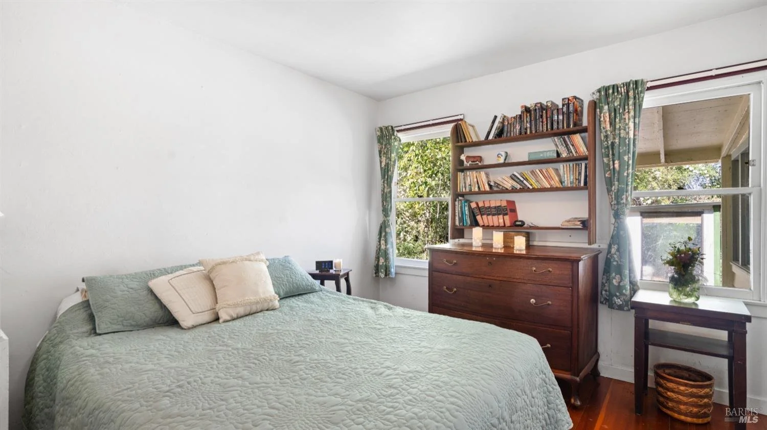 Bedroom with a bed, pillows, nightstand, wooden dresser with a bookshelf on top, two windows with curtains, and a small table with a flower vase.