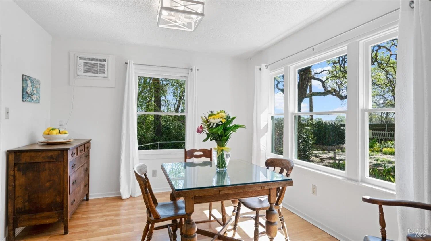 Dining room with wooden table and four chairs, vase with flowers on the table, large windows with white curtains showing trees outside, wooden floor, white walls, and a ceiling light fixture.