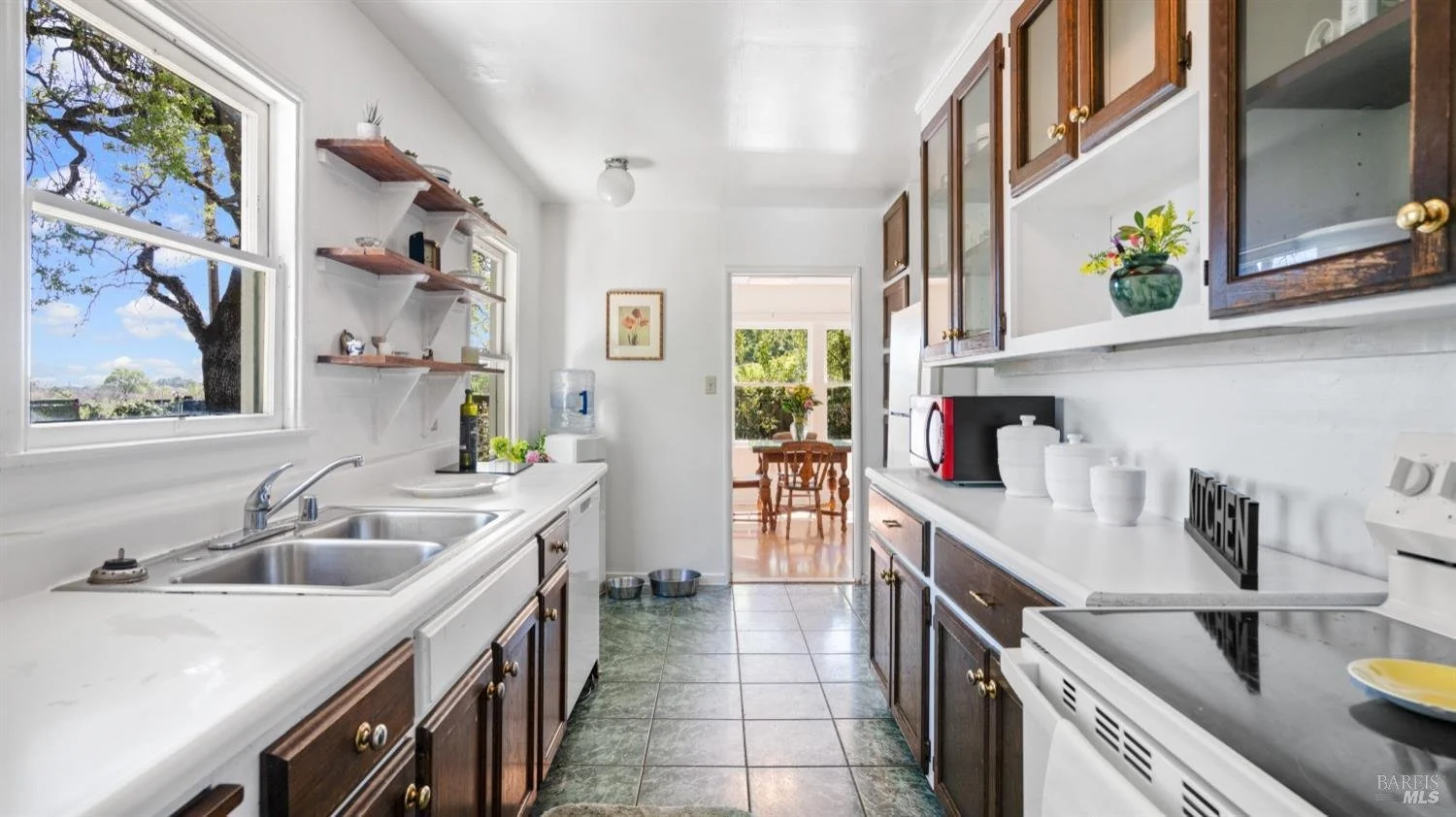 Kitchen with white countertops, wooden cabinets with glass doors, a double sink, open shelving, and a window overlooking a tree. Through a doorway, there is a dining area with a wooden table and chairs, and sunlight coming in.