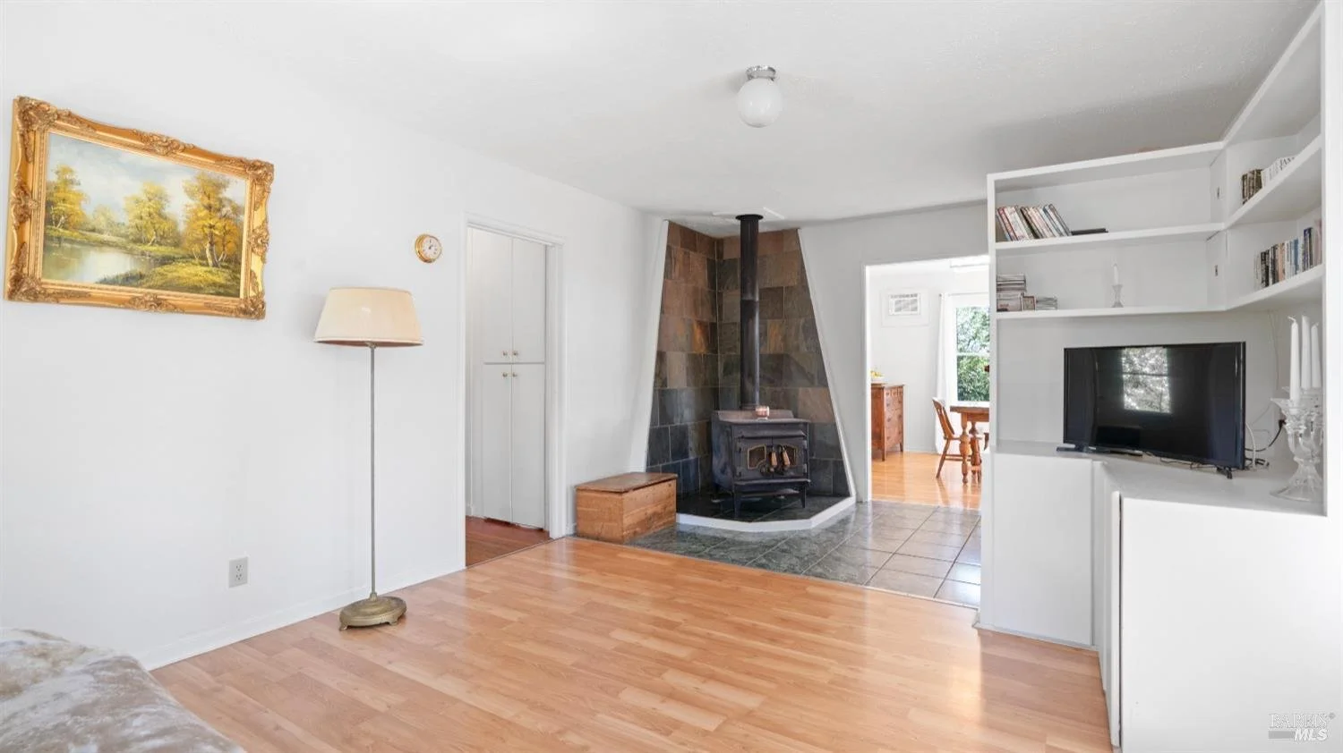 Living room with white walls, a gold-framed landscape painting, a floor lamp, a wood stove with a chimney on a tiled corner, and a white bookshelf with a flat-screen TV. A doorway leads to a dining area with a window, wooden table, and chairs.