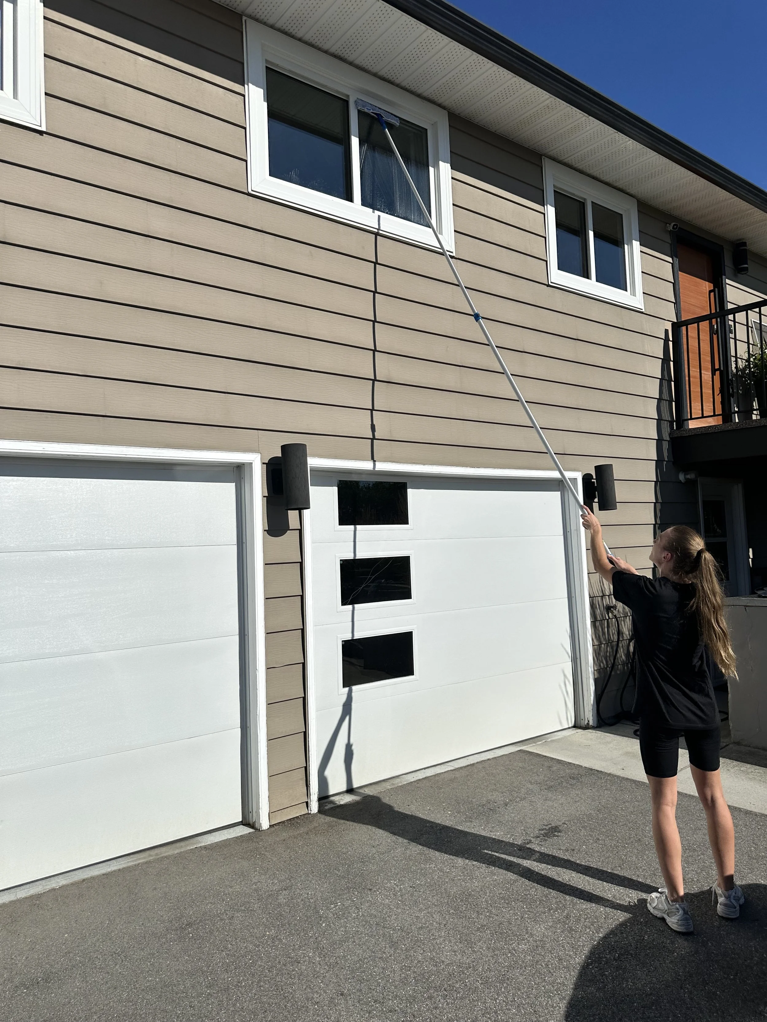 A woman cleaning the upper window of a house with a long extendable squeegee on a sunny day.
