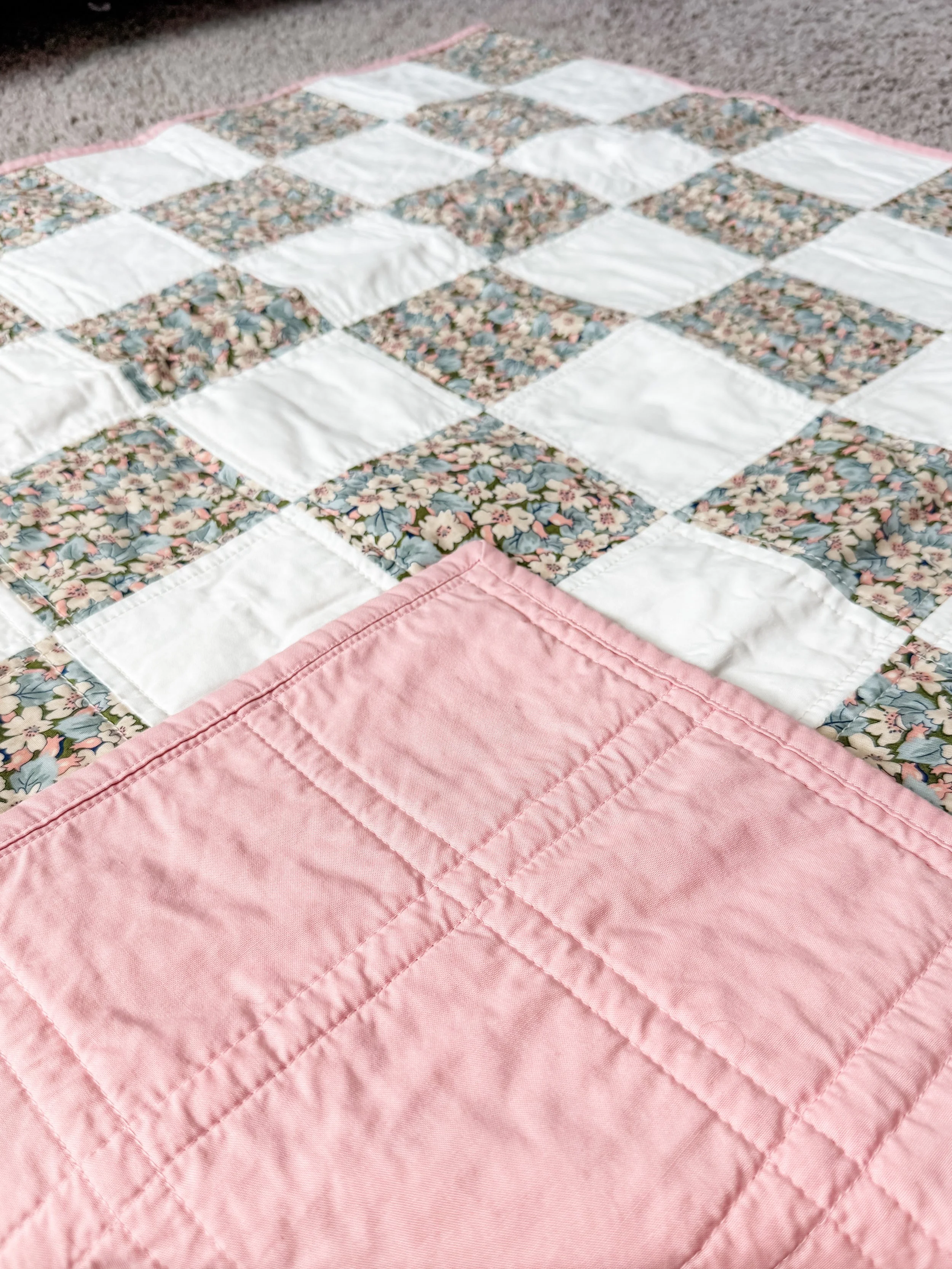 Close-up of a quilt with pink and floral patterns, resting on a carpeted floor.
