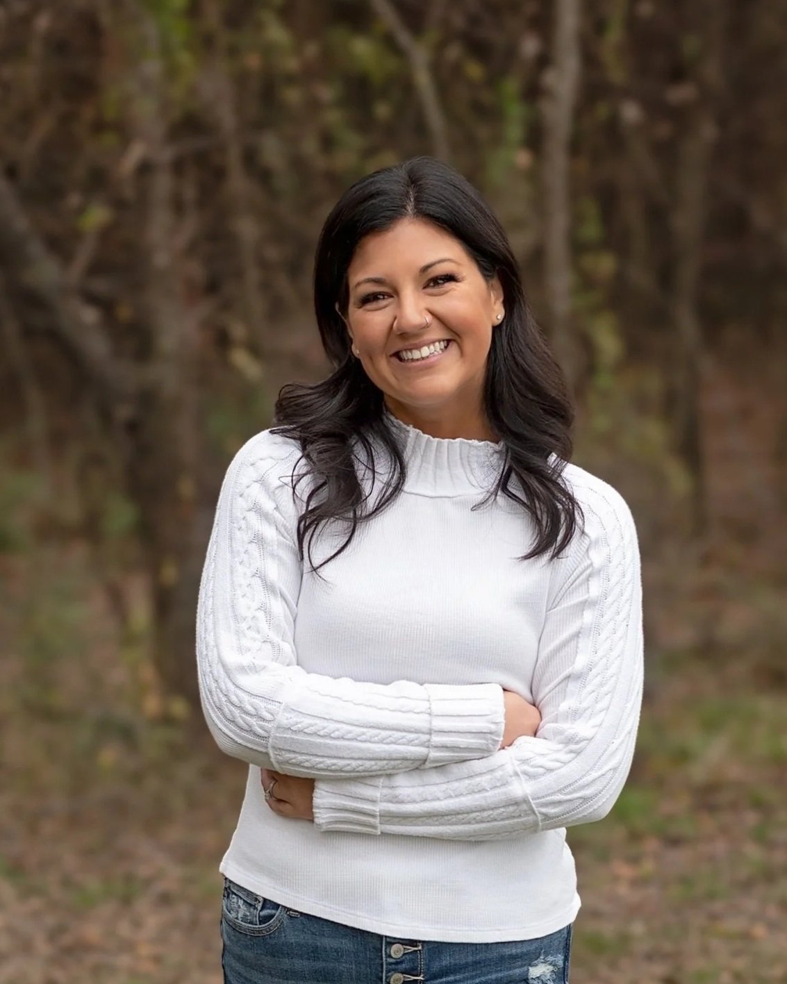 A woman standing outdoors with a blurred natural background, smiling, wearing a white sweater and blue jeans.