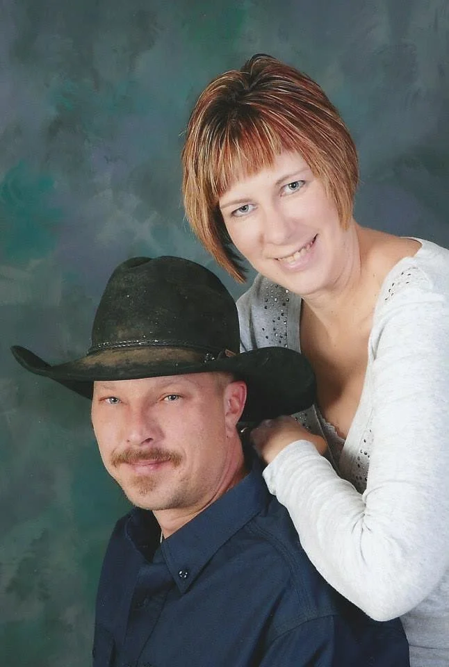 A woman with short reddish hair and a man wearing a cowboy hat and a dark button-up shirt pose together against a painted backdrop.