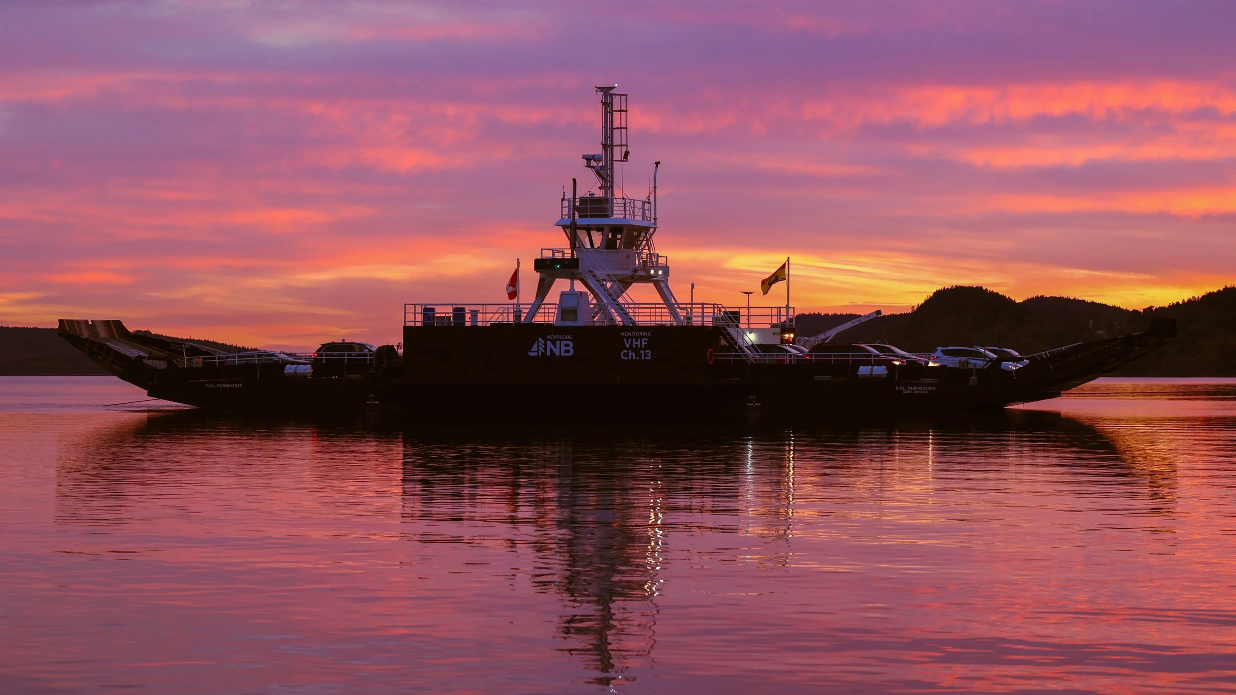 A large blue and black research vessel floating on calm water during a vibrant sunset with pink, orange, and purple sky, and distant hills in the background.