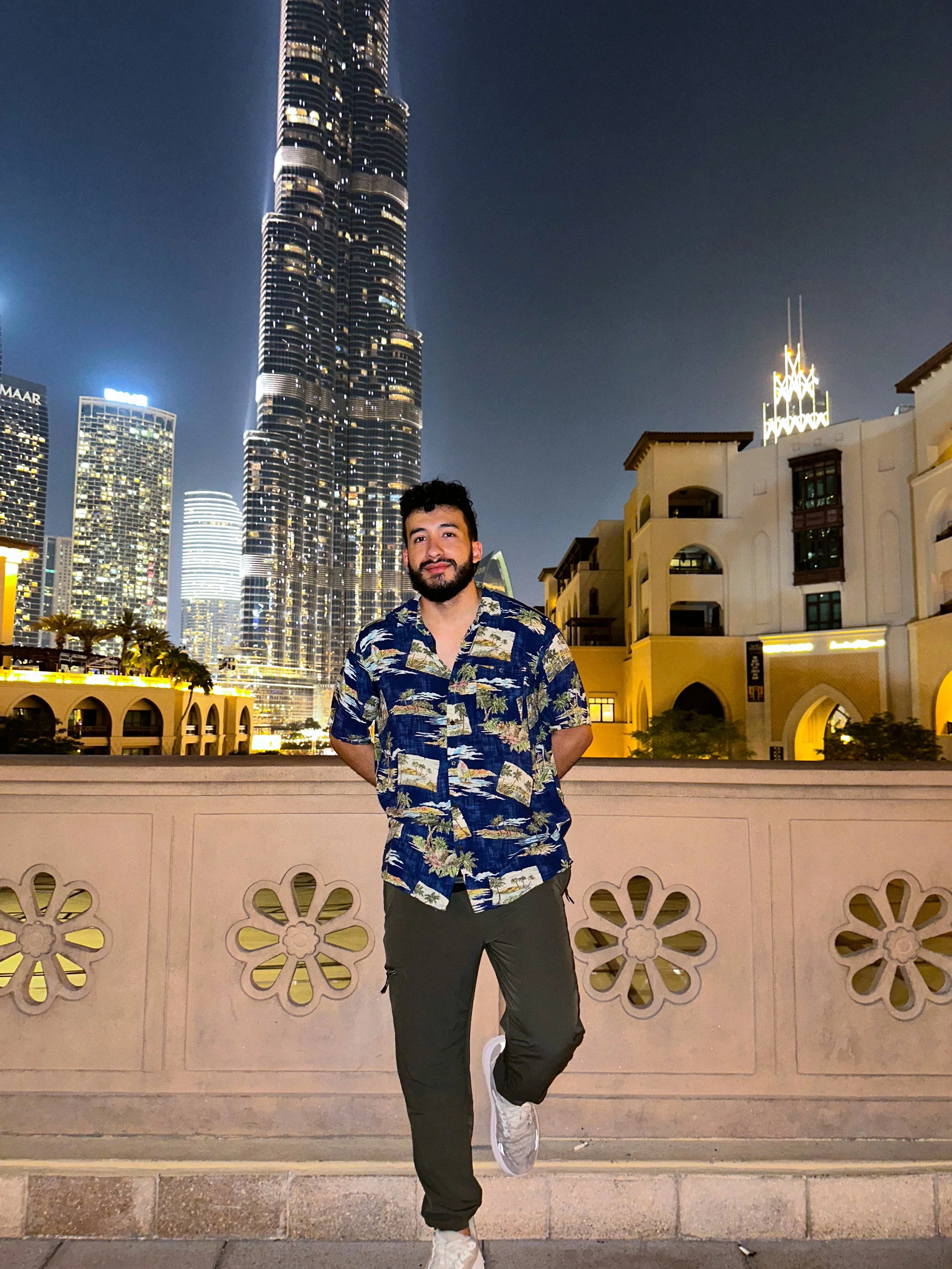A young man with a beard and dark curly hair is standing outdoors at night in front of a decorative wall, posing with one leg bent and hands in his pockets. He is wearing a Hawaiian-style shirt with a floral pattern, black pants, and white sneakers. Behind him is a cityscape with tall illuminated skyscrapers, including the Burj Khalifa, in Dubai.