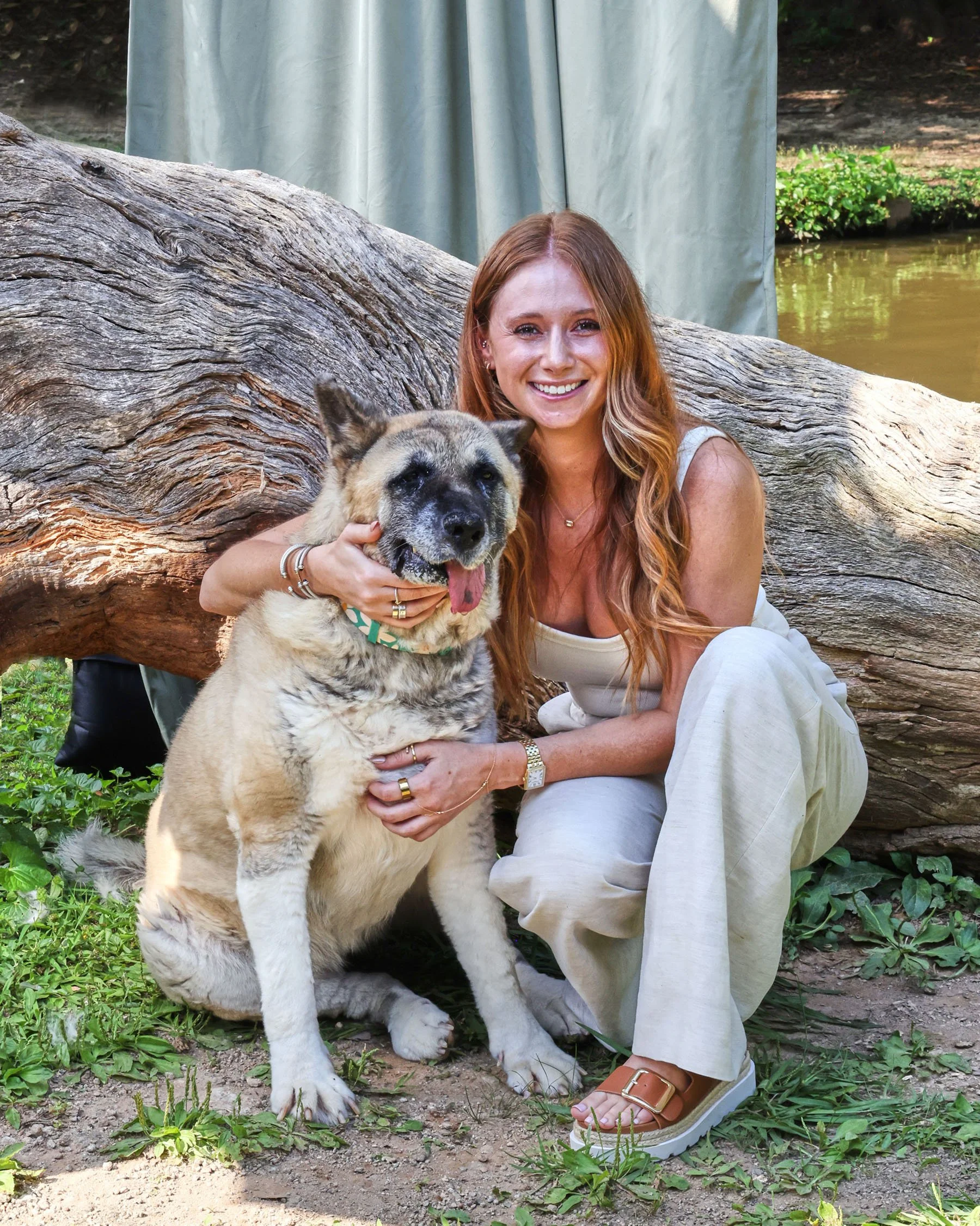 Woman with red hair, sitting next to dog with her tongue out, leaning against a log.