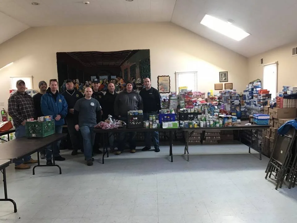 Group of people standing behind tables filled with food and supplies, with numerous boxes of canned and packaged goods stacked on tables and the floor.