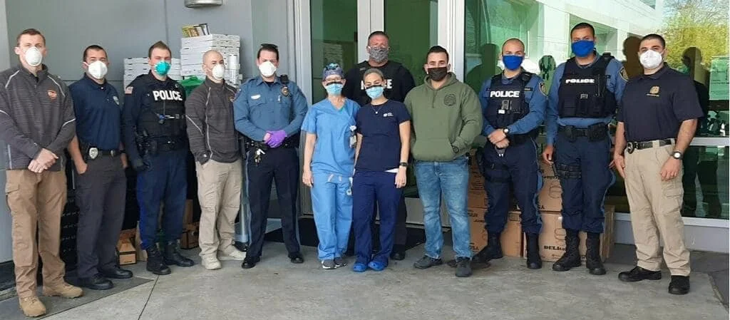 Group of police officers, healthcare workers, and civilians wearing masks standing outdoors in front of a glass building.