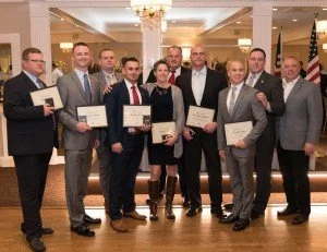 A group of nine men and women dressed in formal attire standing together in a banquet hall, holding certificates or awards, smiling for the photo.