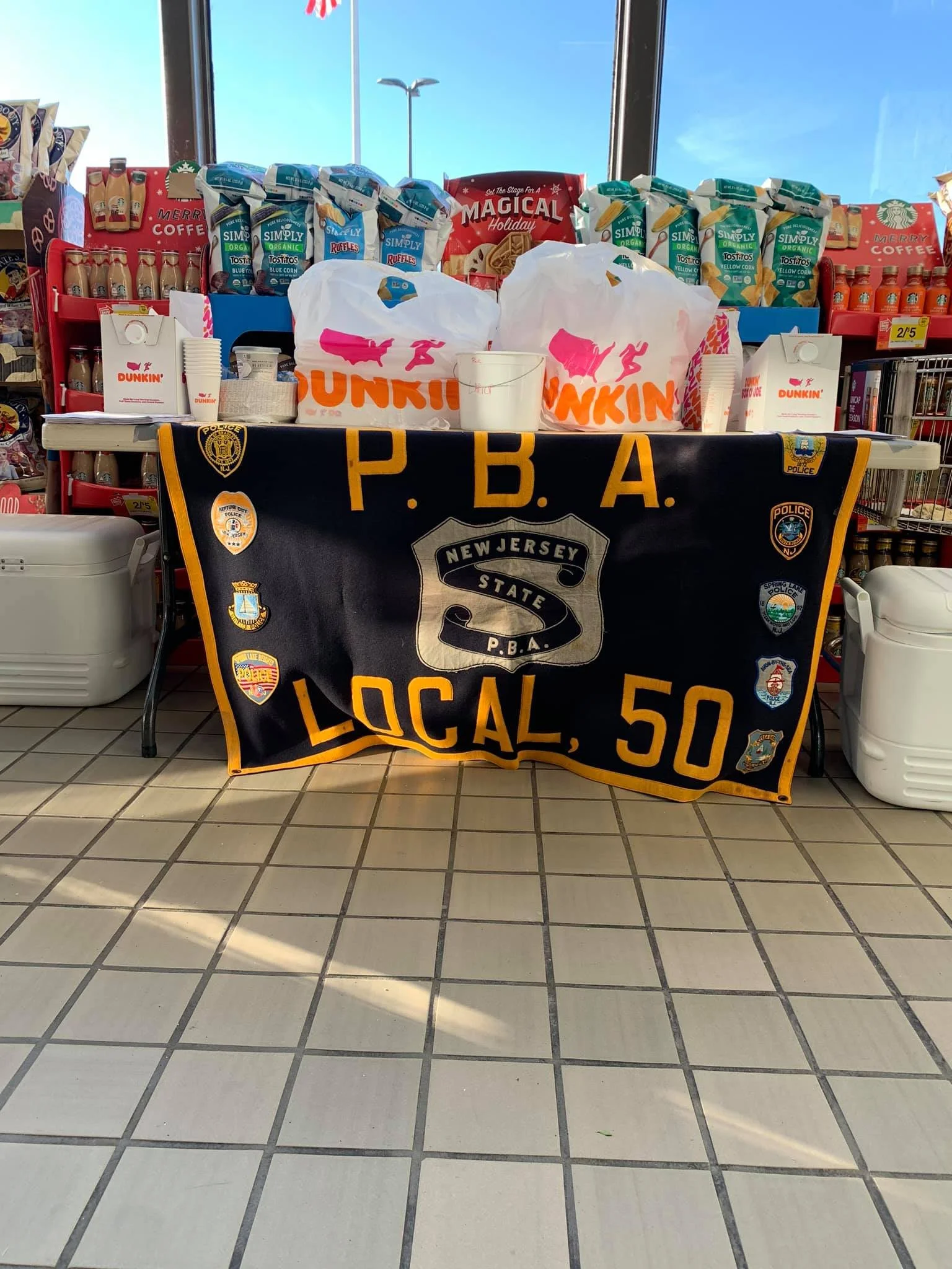 Table with Dunkin' Donuts bags and coffee cups, police banner from New Jersey State Police Local 50, and Organization of Police badges in a store.
