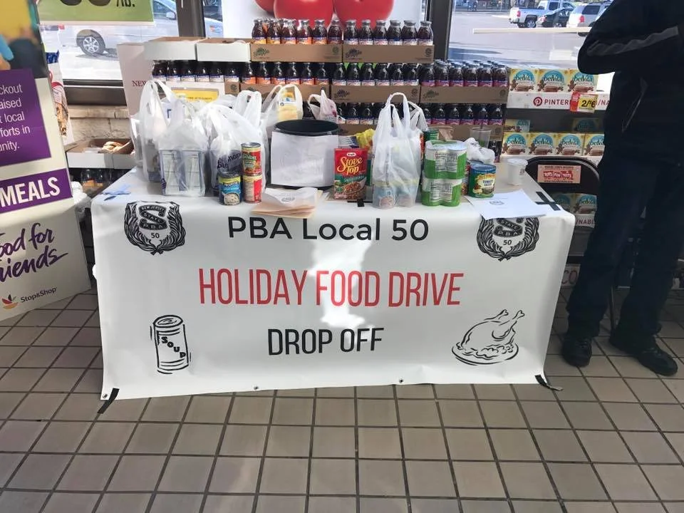Table at a holiday food drive organized by PBA Local 50, with canned and packaged food items for donation.
