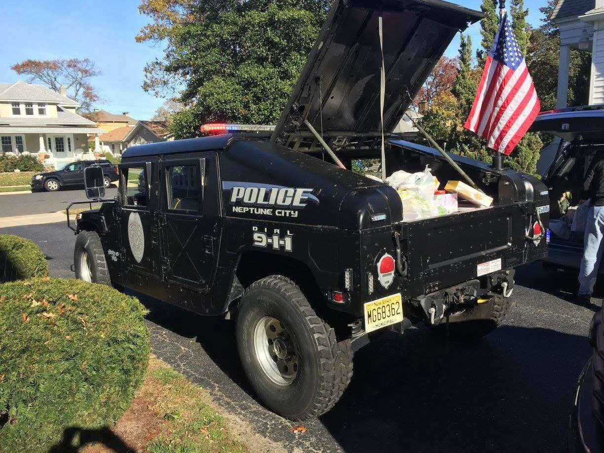 A black police vehicle with its rear open, containing supplies and a small American flag, parked outdoors in a residential neighborhood with trees and houses in the background.
