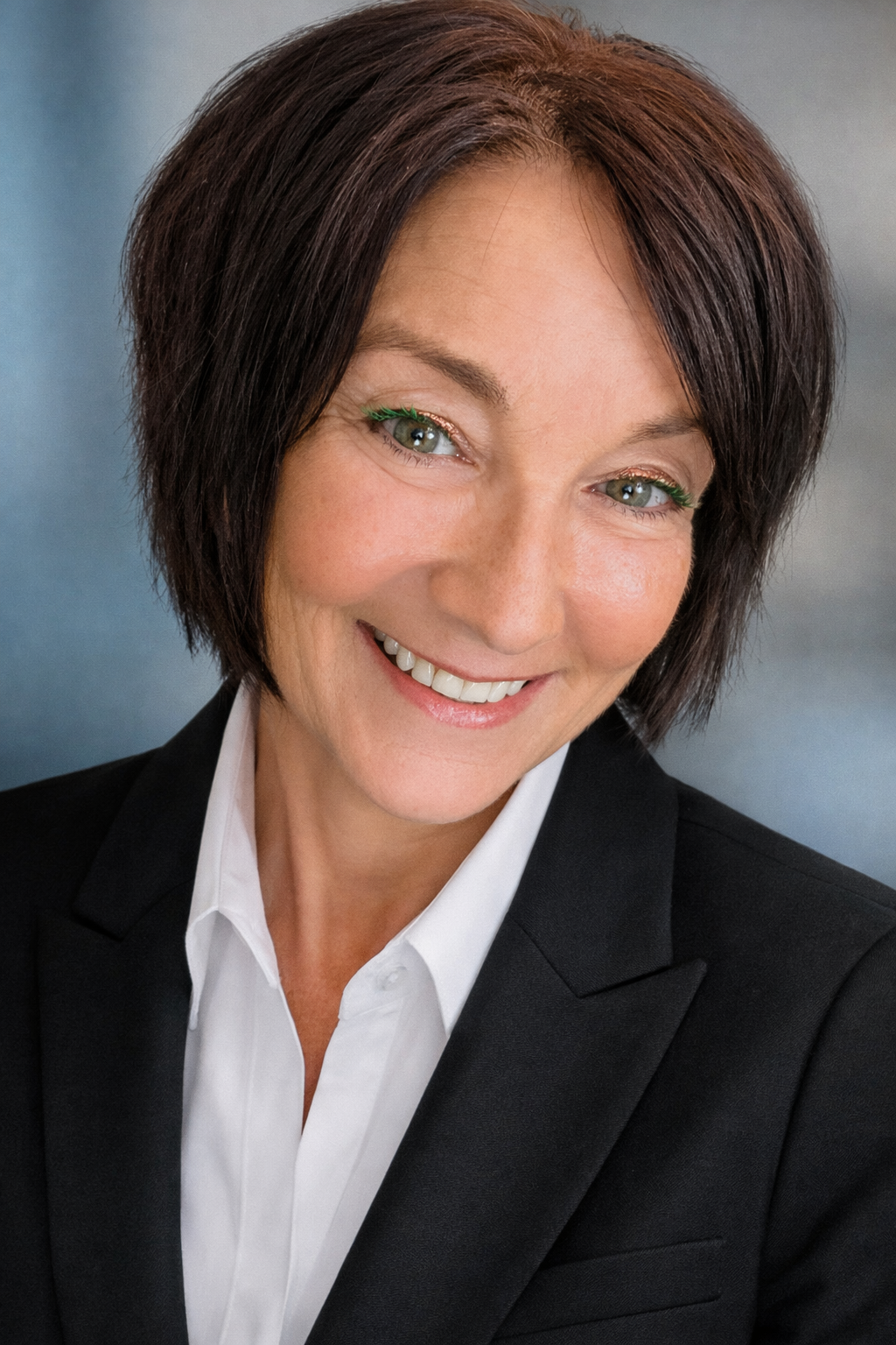 A woman with shoulder-length dark brown hair, light skin, and green eyes, wearing a black blazer and white collared shirt, smiling against a gradient background.