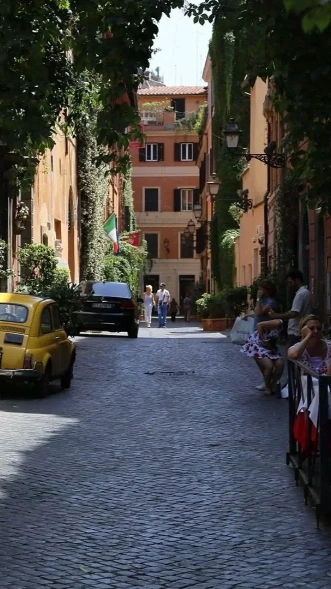 Cobblestone street in Rome with historic buildings, greenery, outdoor dining, and everyday life.