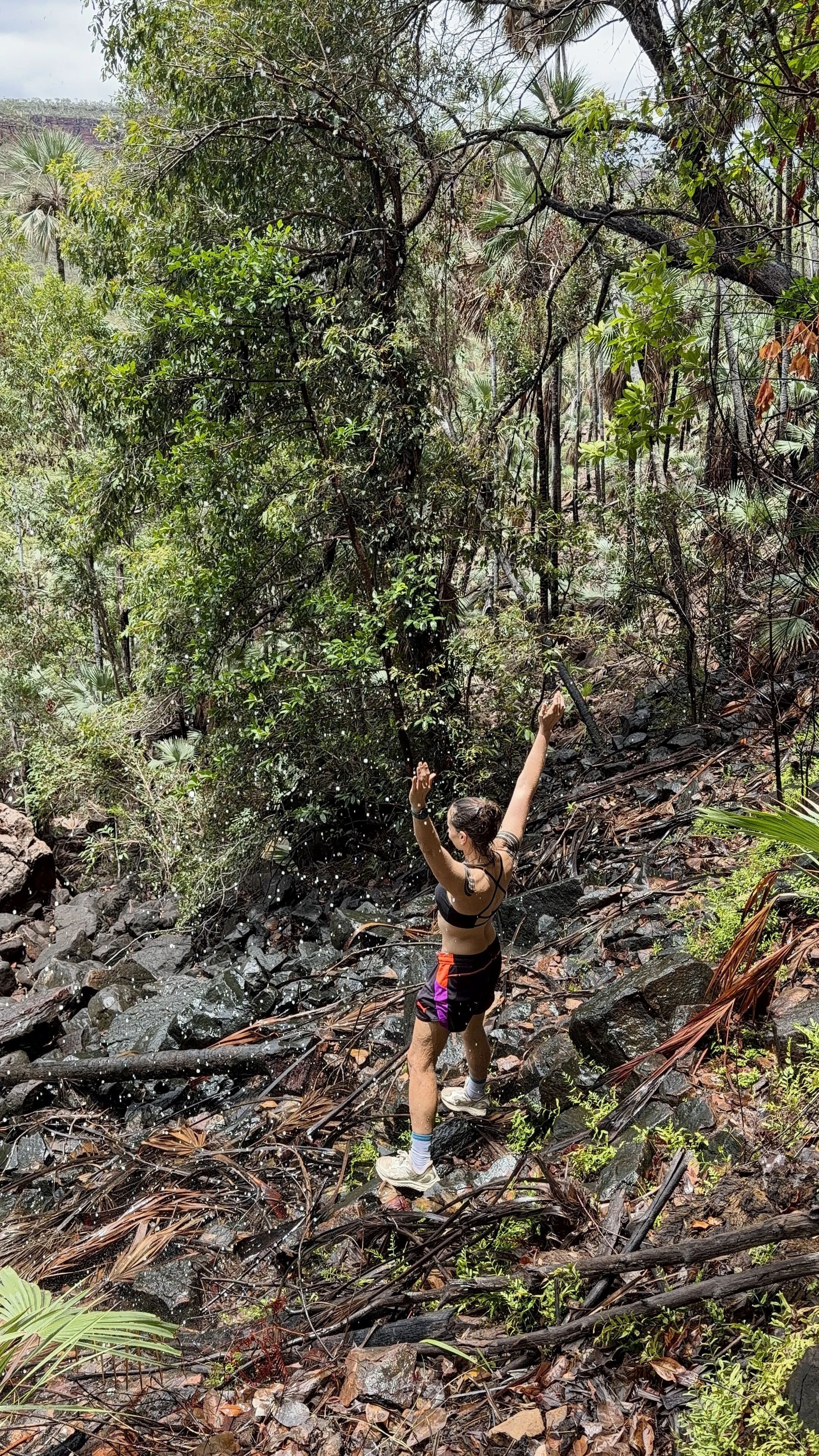 A woman in sportswear standing on wet rocks in a lush, green forest, with arms raised and surrounded by dense foliage.