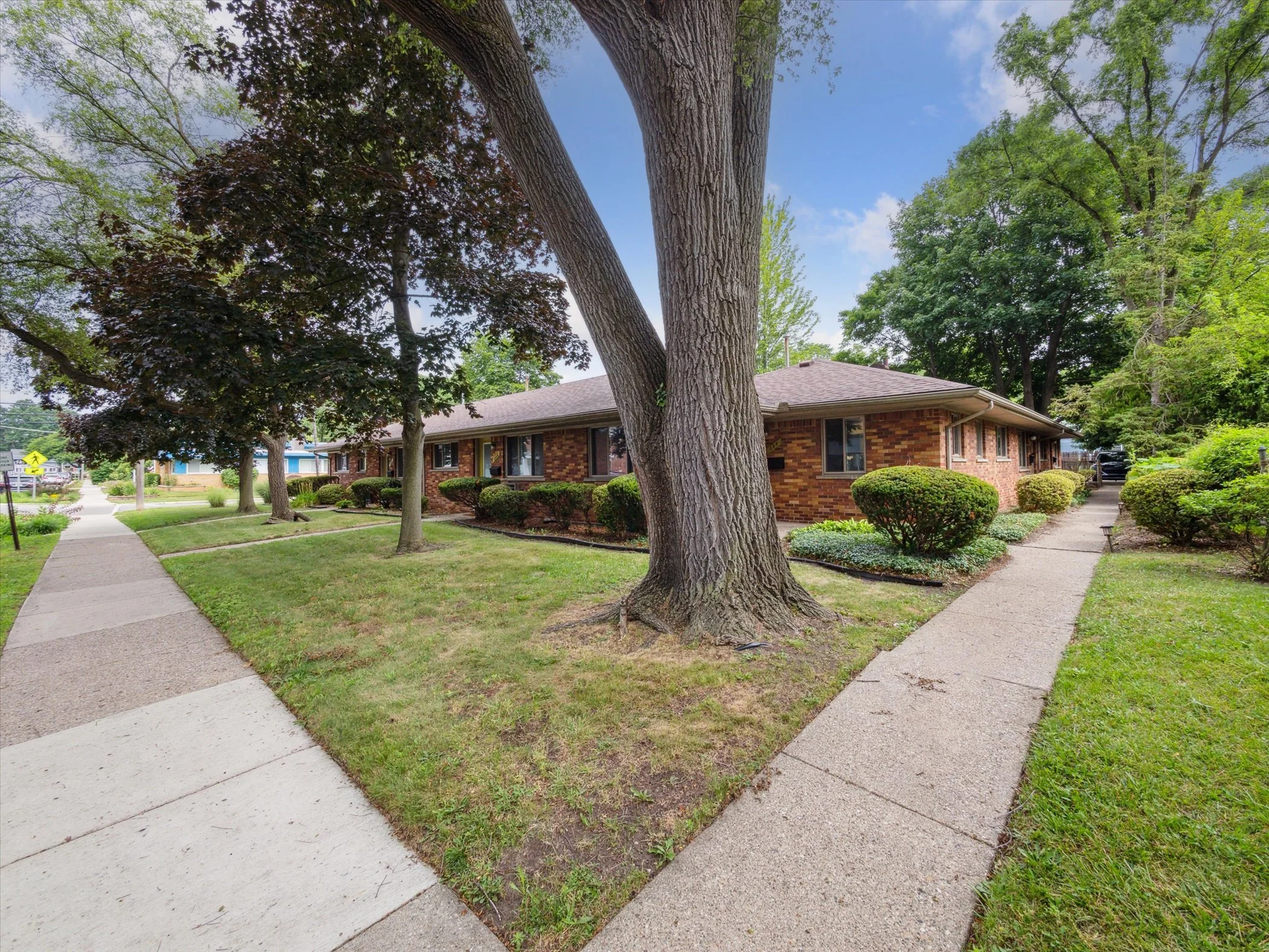 Sidewalks and well-maintained yard with trees and bushes in front of a brick residential building amid lush green trees under a partly cloudy sky.