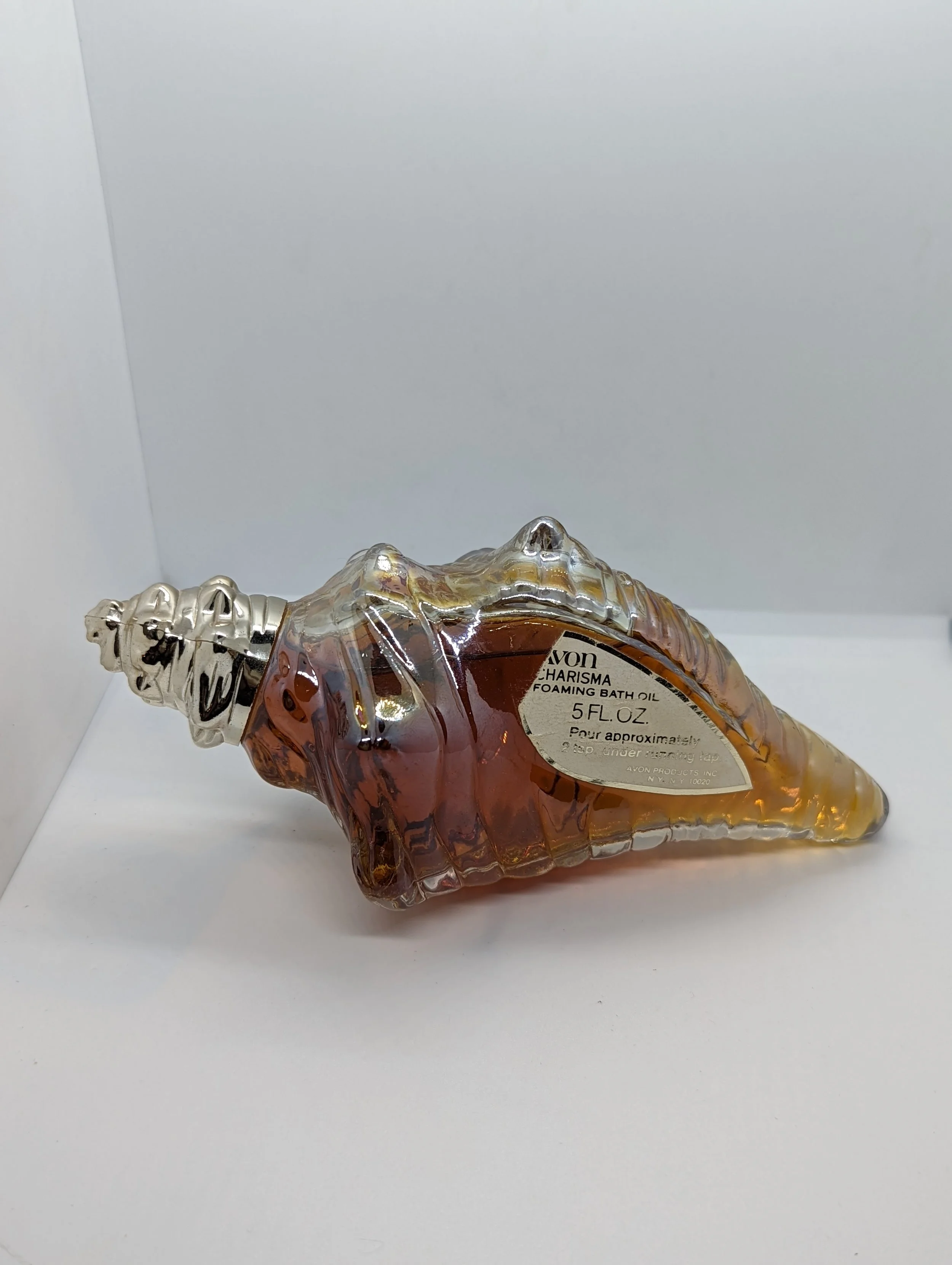A seashell-shaped glass bottle filled with amber-colored foaming bath oil lying on its side on a white surface.