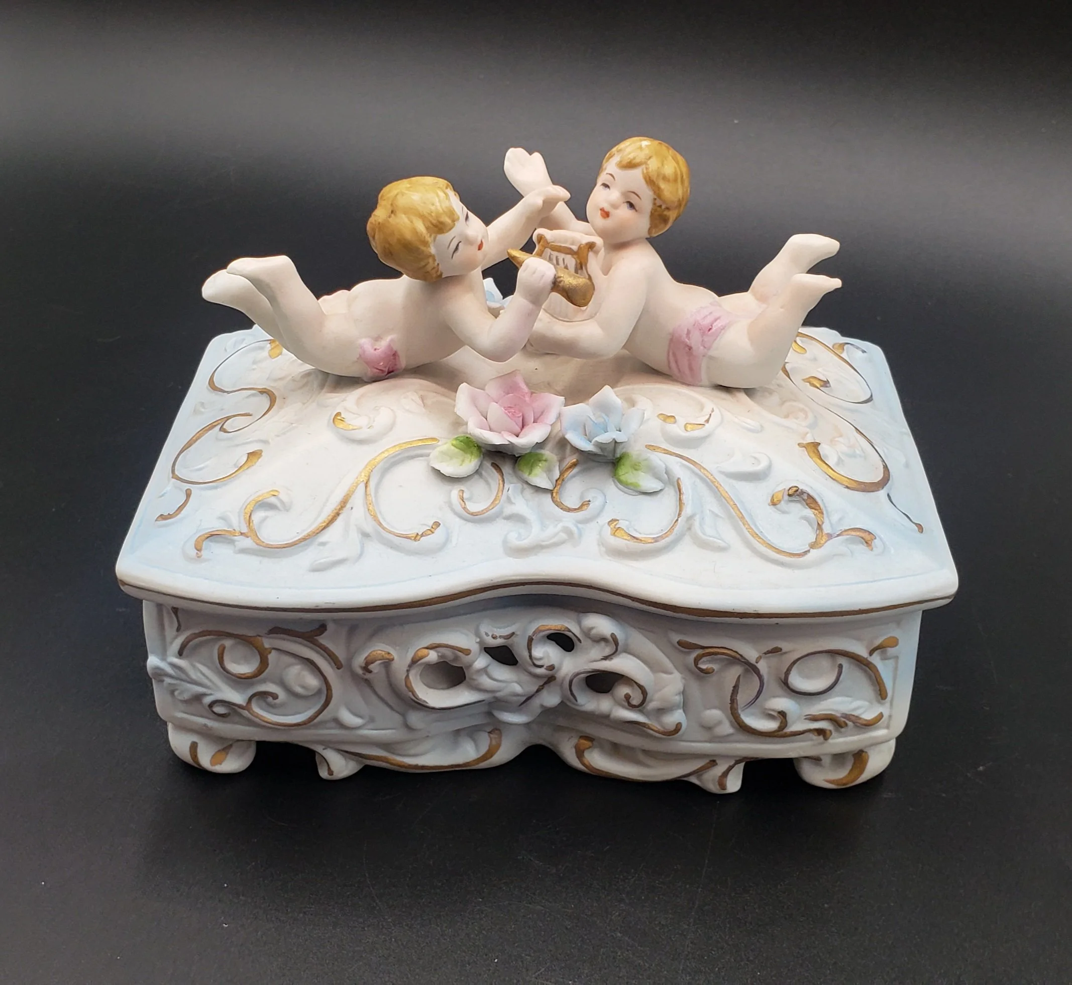 Decorative porcelain box with two cherubs playing musical instruments on top, surrounded by pink and blue flowers, and gold accents.