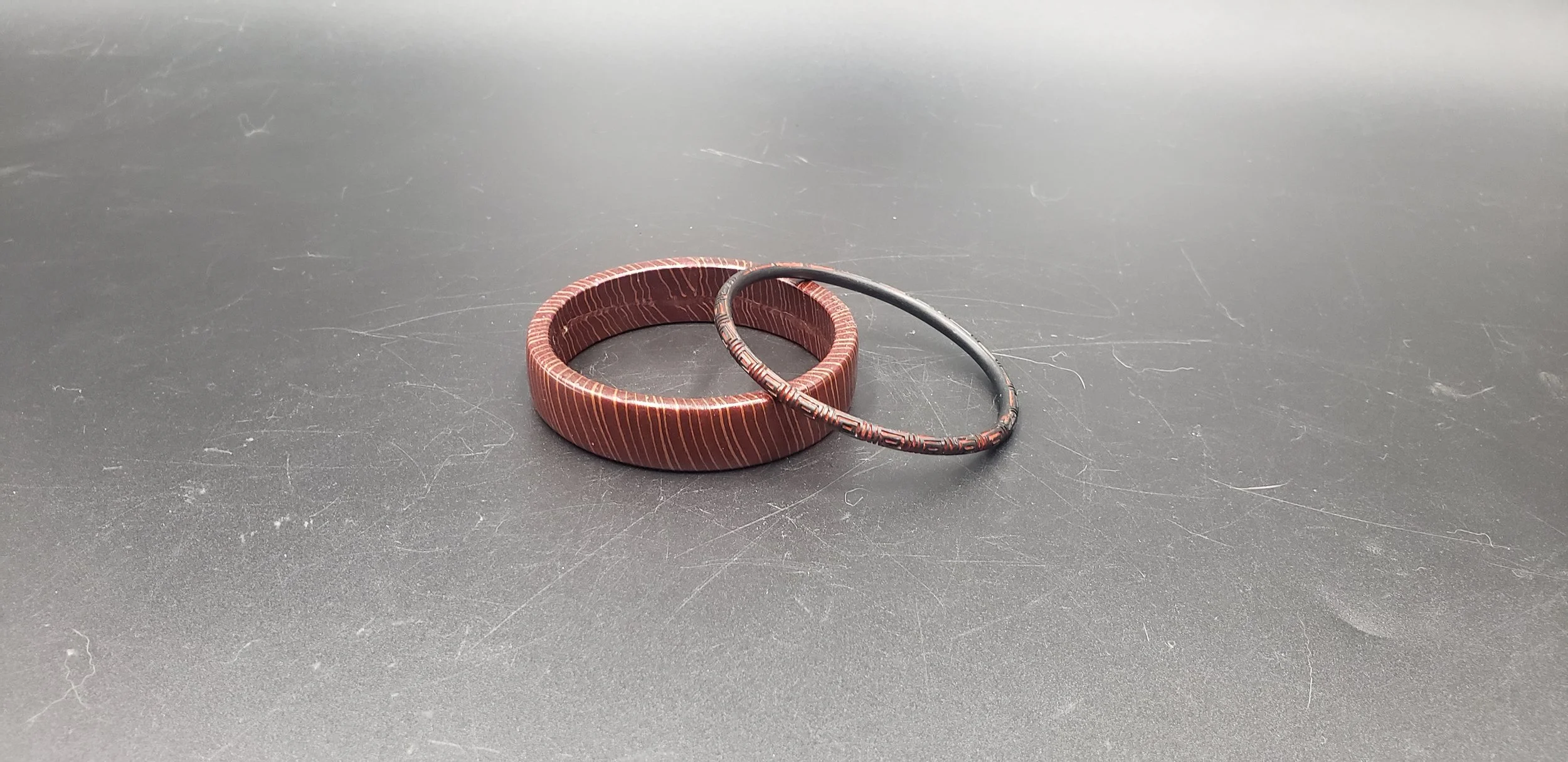 A brown bangle bracelet with a striped pattern and a black circular bracelet with brown accents on a scratched gray surface.