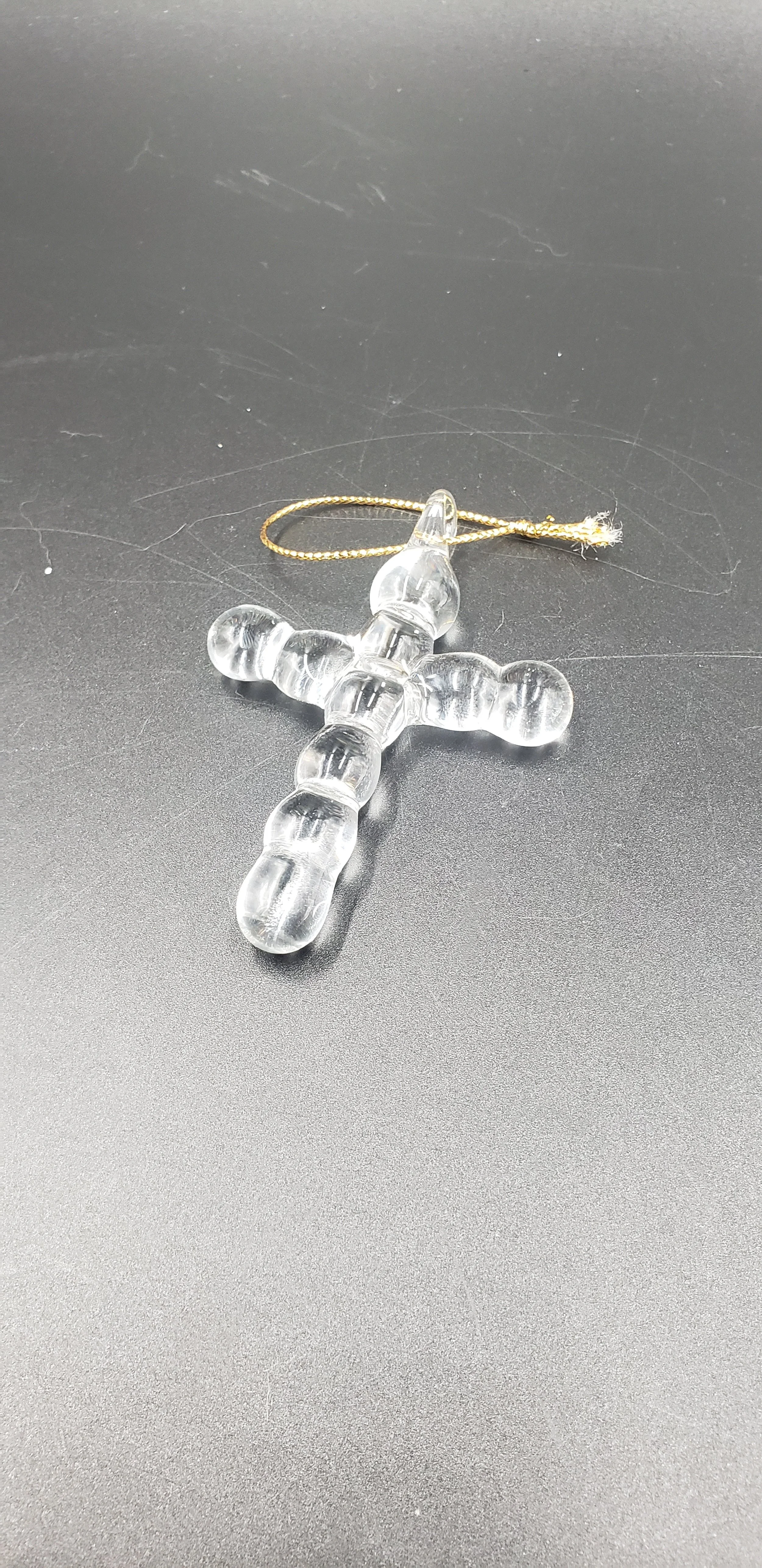 A clear glass or plastic cross-shaped ornament with a gold thread for hanging, resting on a dark surface.