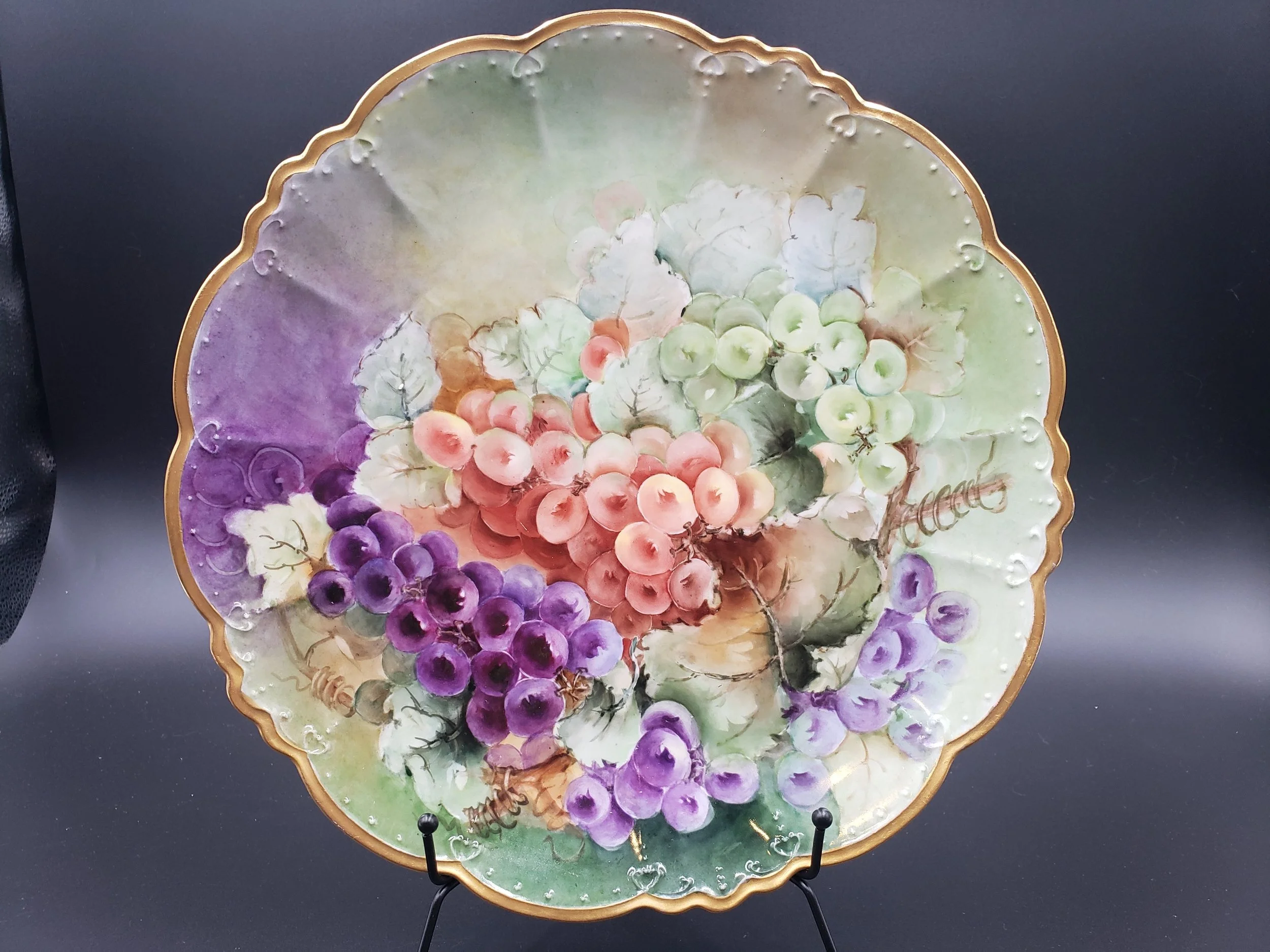 Decorative porcelain plate with painted clusters of purple, pink, and green grapes, edged with a gold scalloped border, displayed on a stand against a dark background.