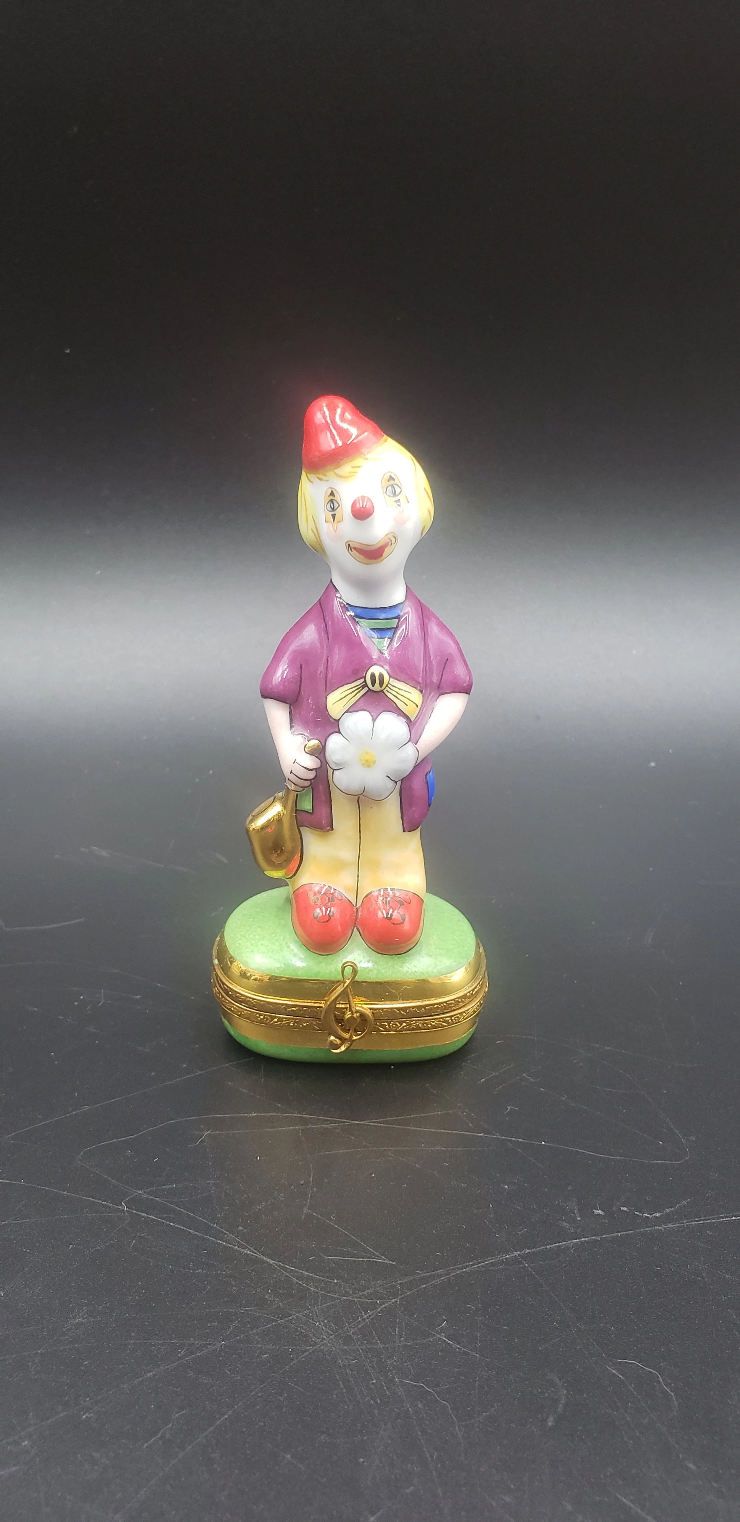 Colorful clown figurine with a red hat, yellow hair, purple shirt, yellow pants, red shoes, and a white flower, standing on a decorated green and gold base.