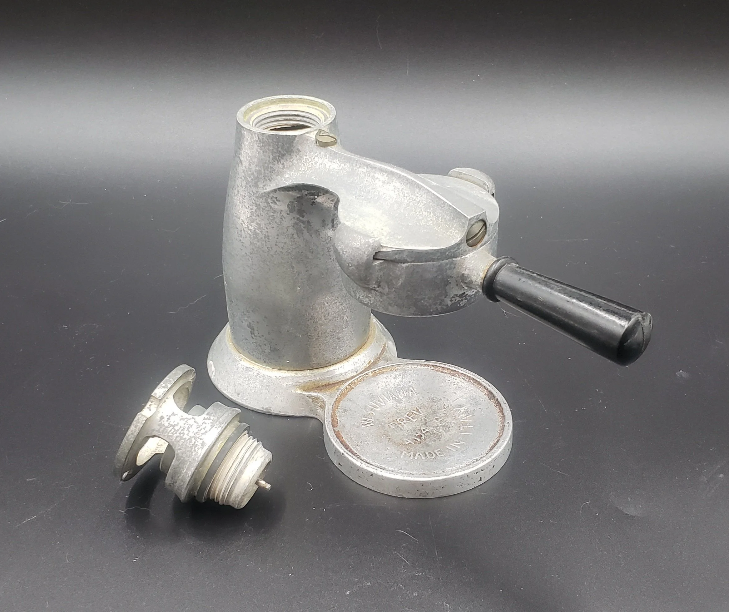 Metal valve with black handle and metallic fitting on a black surface, with an attached circular metal plate.