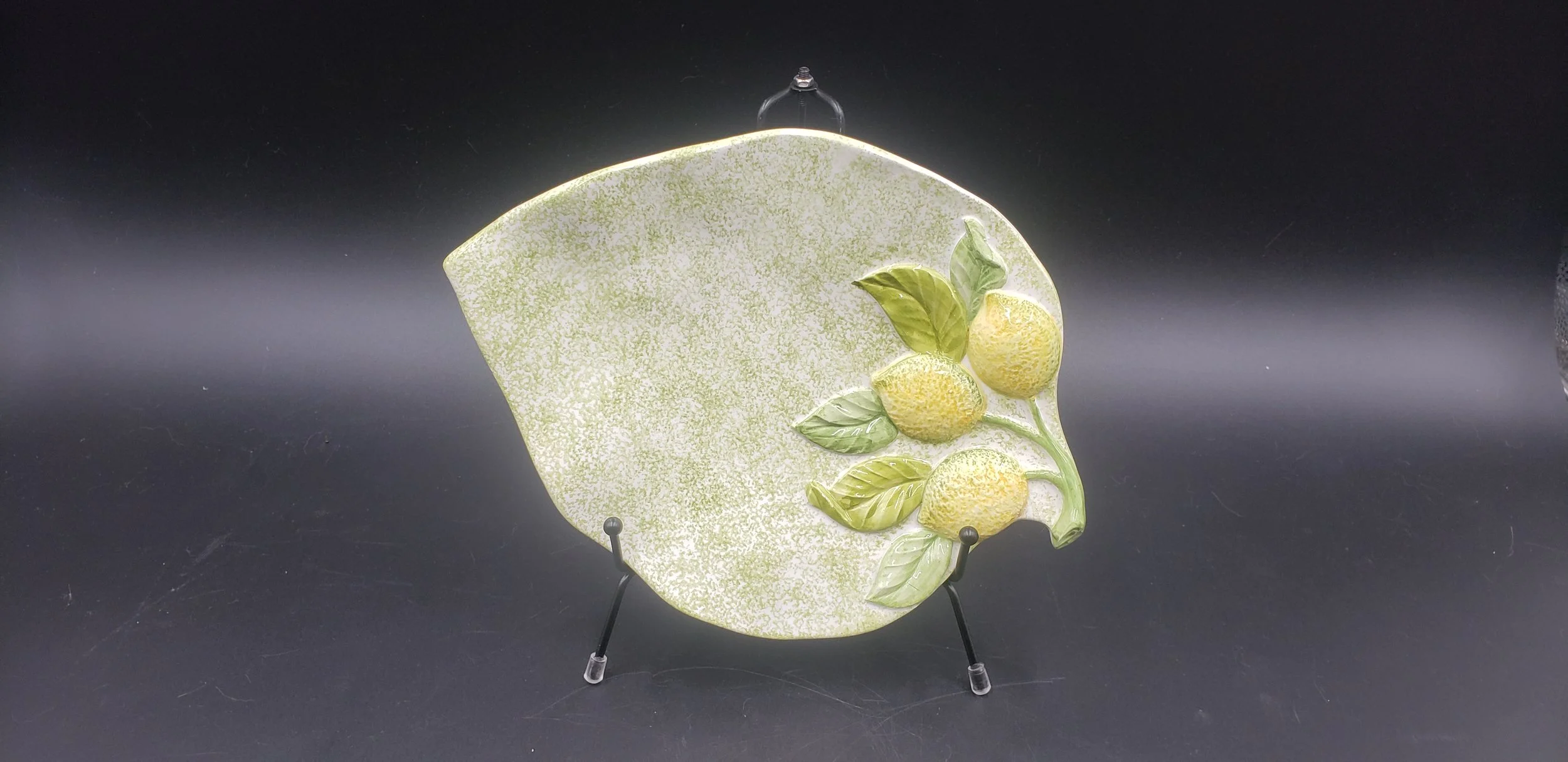 Decorative ceramic plate with lemon and leaves design, displayed on a stand against a black background.
