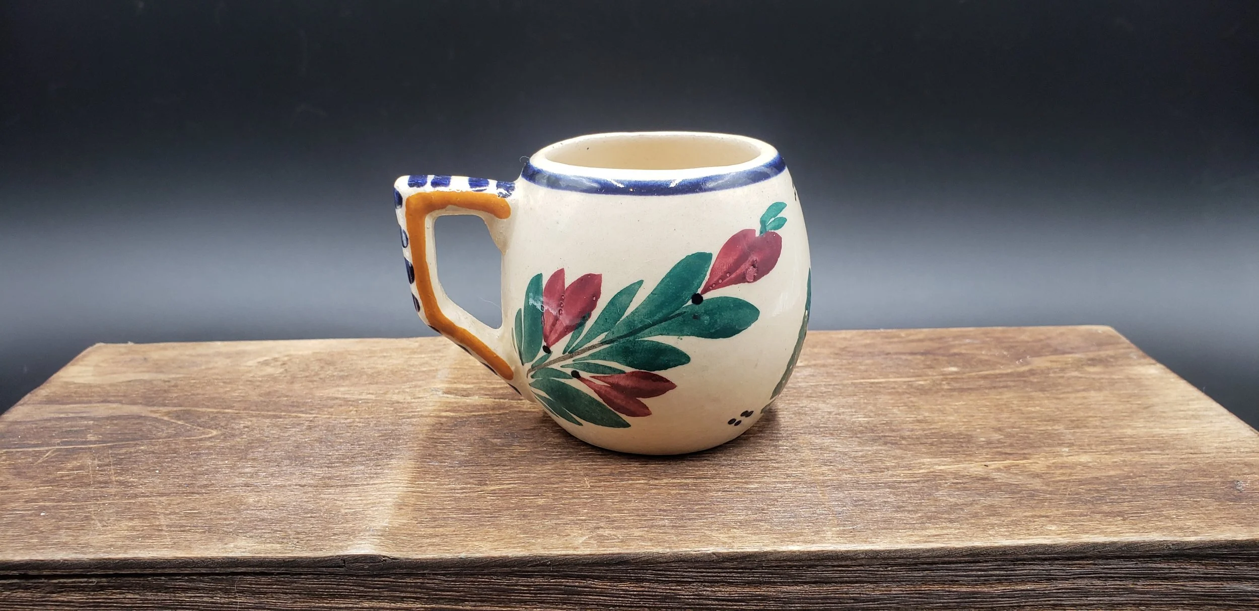 Decorative ceramic mug with a floral design in red and green, handmade style, placed on a wooden surface with a dark gray background.