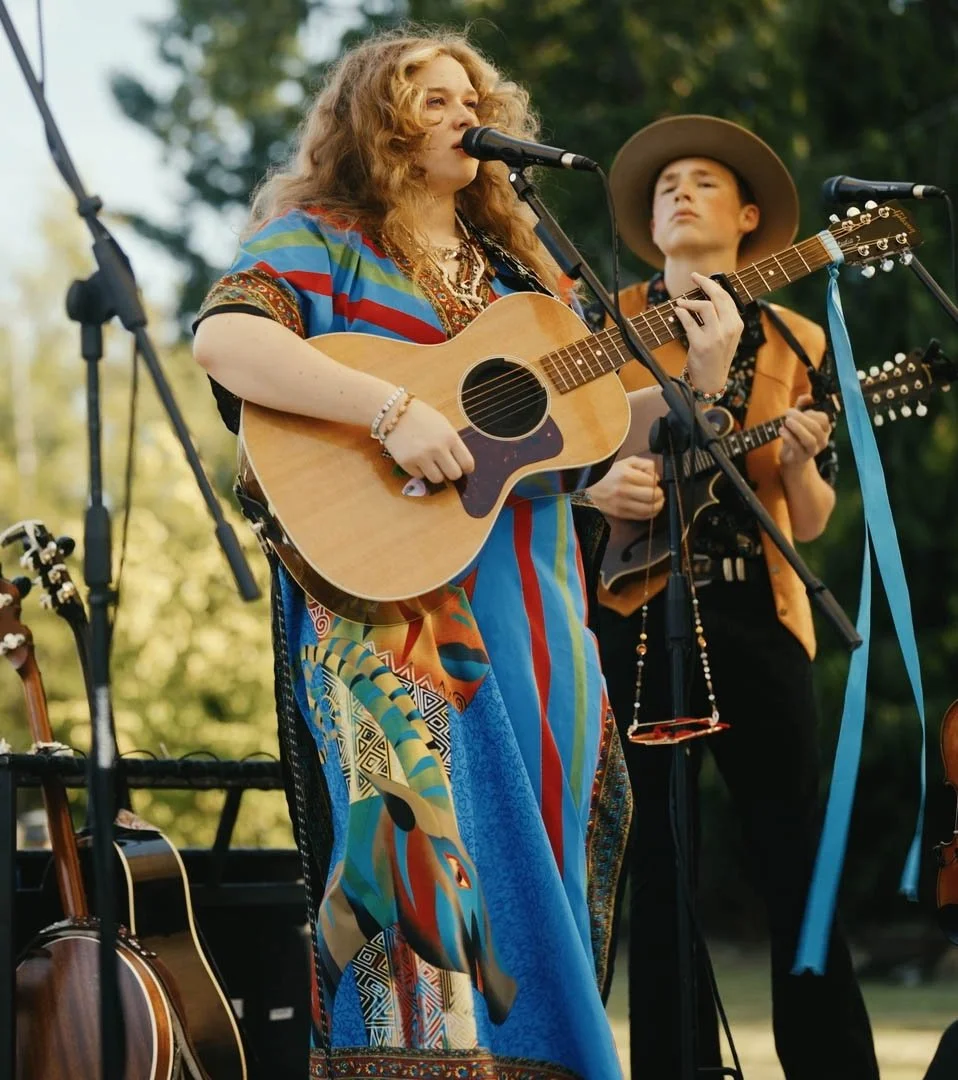 Two women playing guitars and singing on an outdoor stage during the daytime, surrounded by trees.