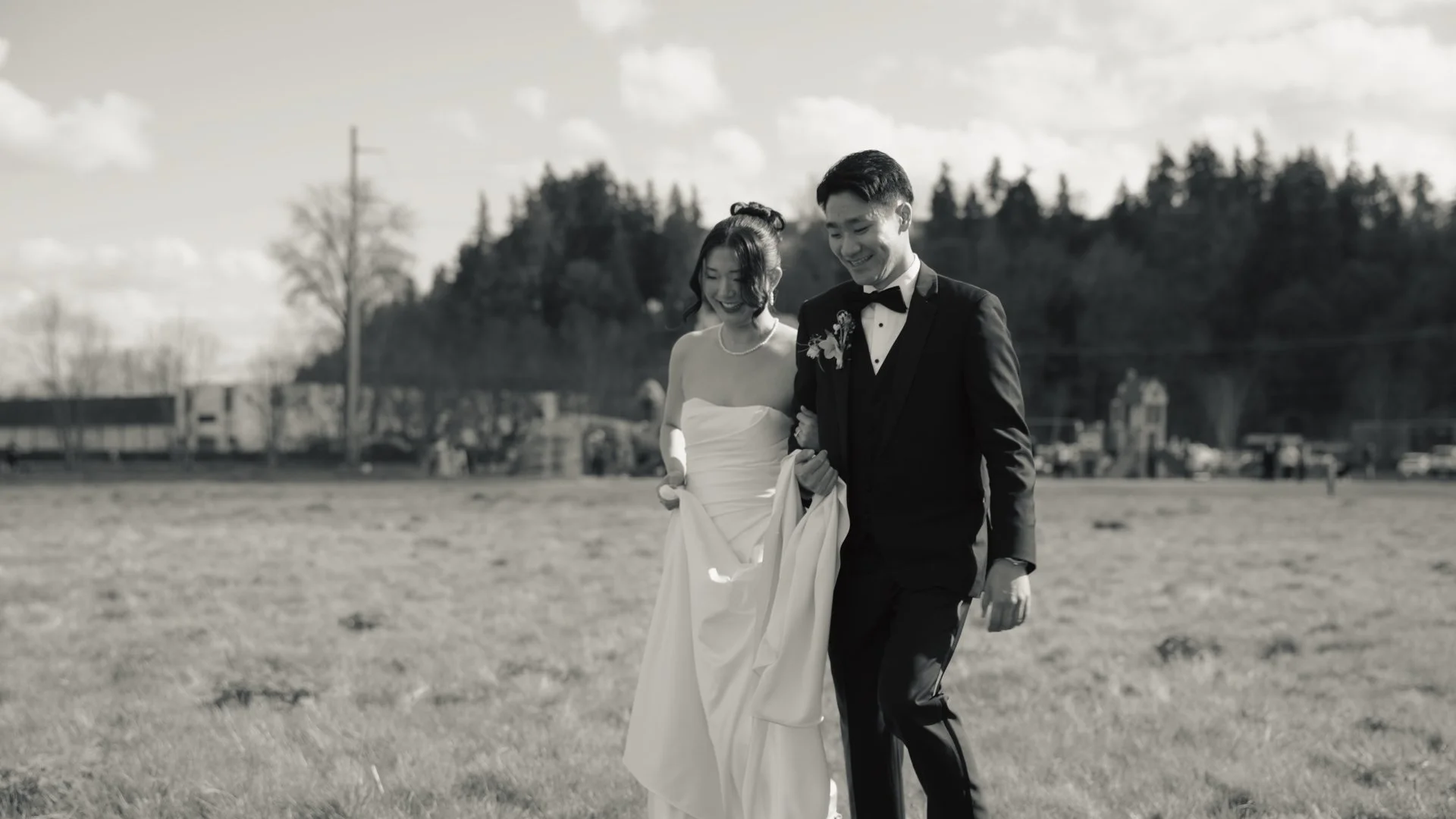 A bride and groom walking together on a grassy field, smiling, with trees and buildings in the background, in black and white. This wedding was at Farm 12 in Puyallup, Washington.