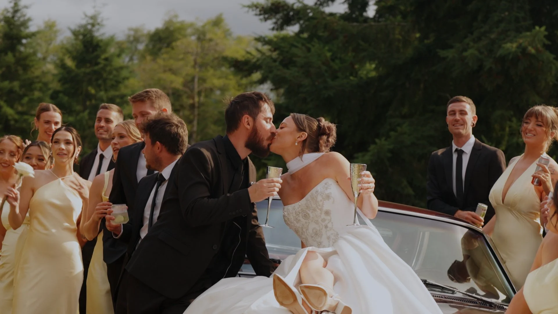 A Washington wedding with a newlywed couple in wedding attire kissing each other, surrounded by wedding guests, outdoors in front of a classic car, with green trees in the background.