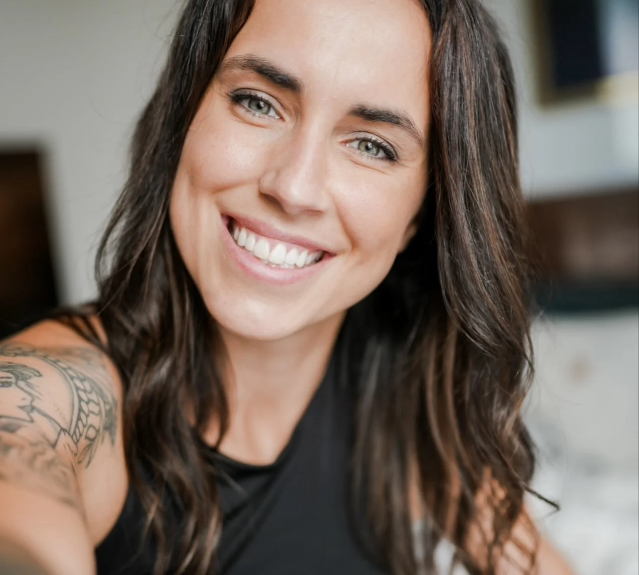 Close-up of a smiling woman with long brown hair, blue eyes, and a tattoo on her shoulder, wearing a black top.
