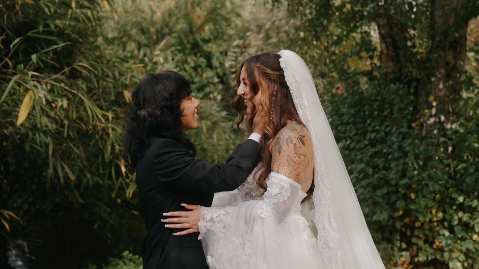 A Seattle wedding with a gay couple, one in a wedding dress and the other in a suit, are embracing outdoors among greenery and bushes, smiling at each other closely.