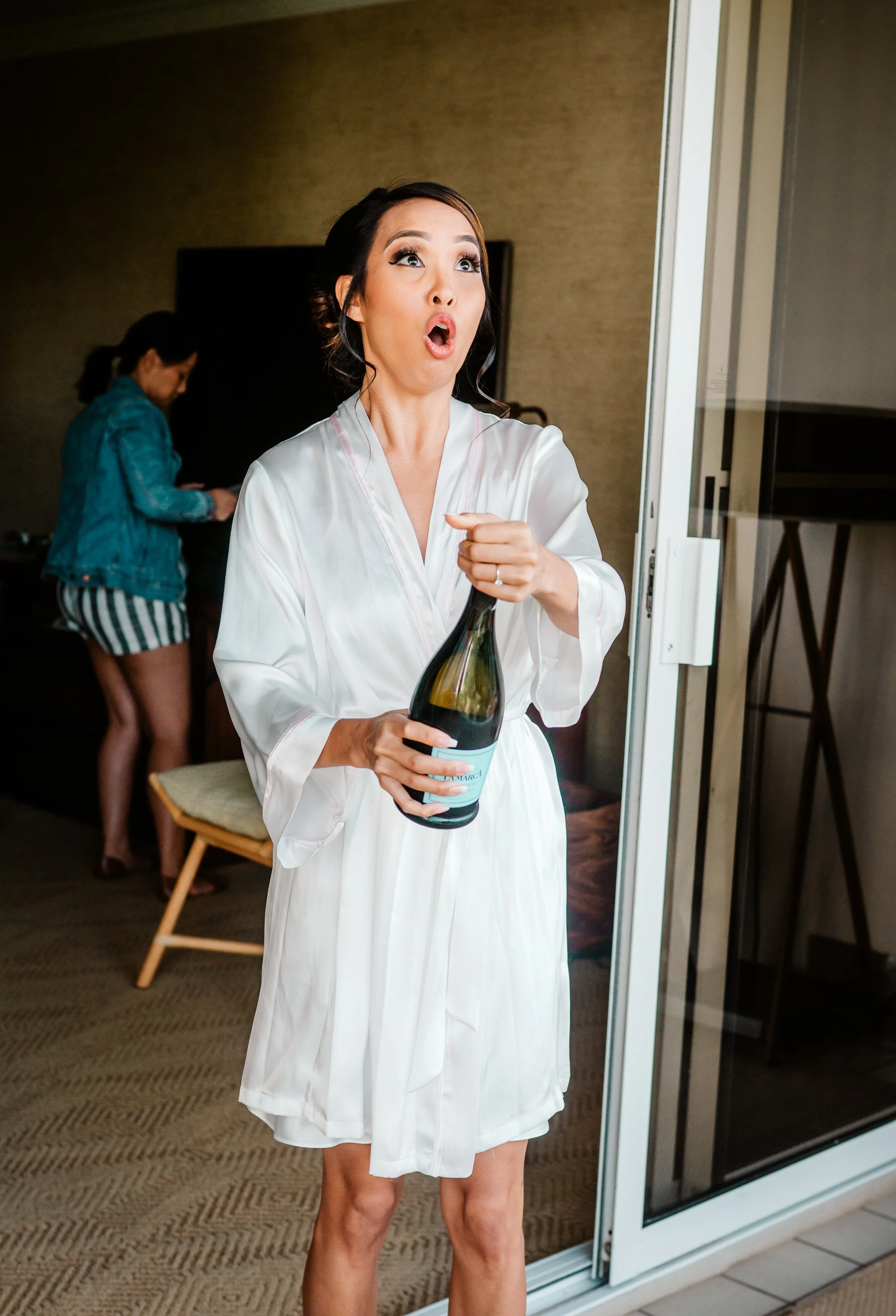 Woman in white robe holding a bottle and reacting with surprise or excitement, standing near a glass door, with another woman in casual clothes in the background.
