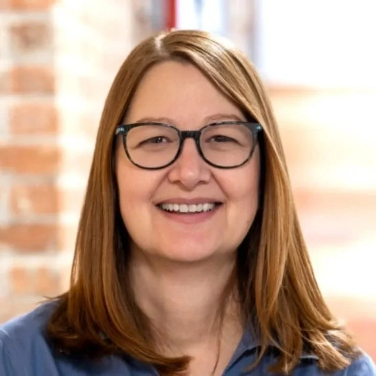 A woman with shoulder-length brown hair wearing glasses and a white collared shirt with an American flag pin, smiling in front of a plain beige background.