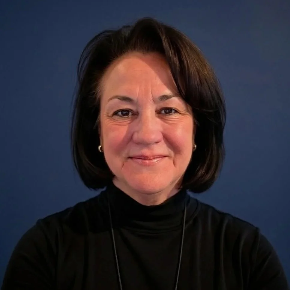 Portrait of a middle-aged woman with gray hair smiling, wearing a black blazer with shimmering details, turquoise jewelry, and a swirl earring, against a plain gray background.