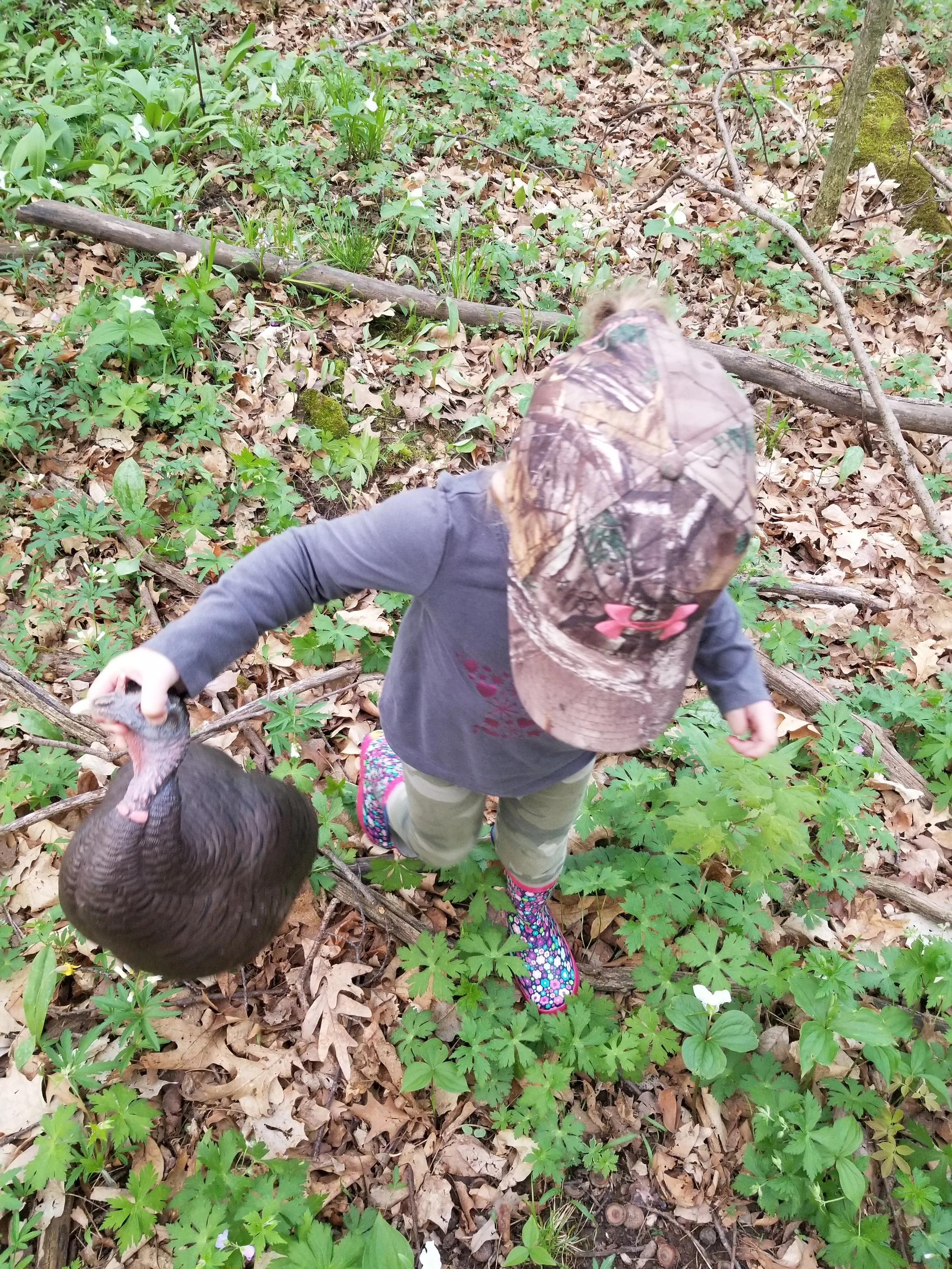 Child wearing a camouflage cap and colorful rain boots in a wooded area holding a wild turkey by its neck.
